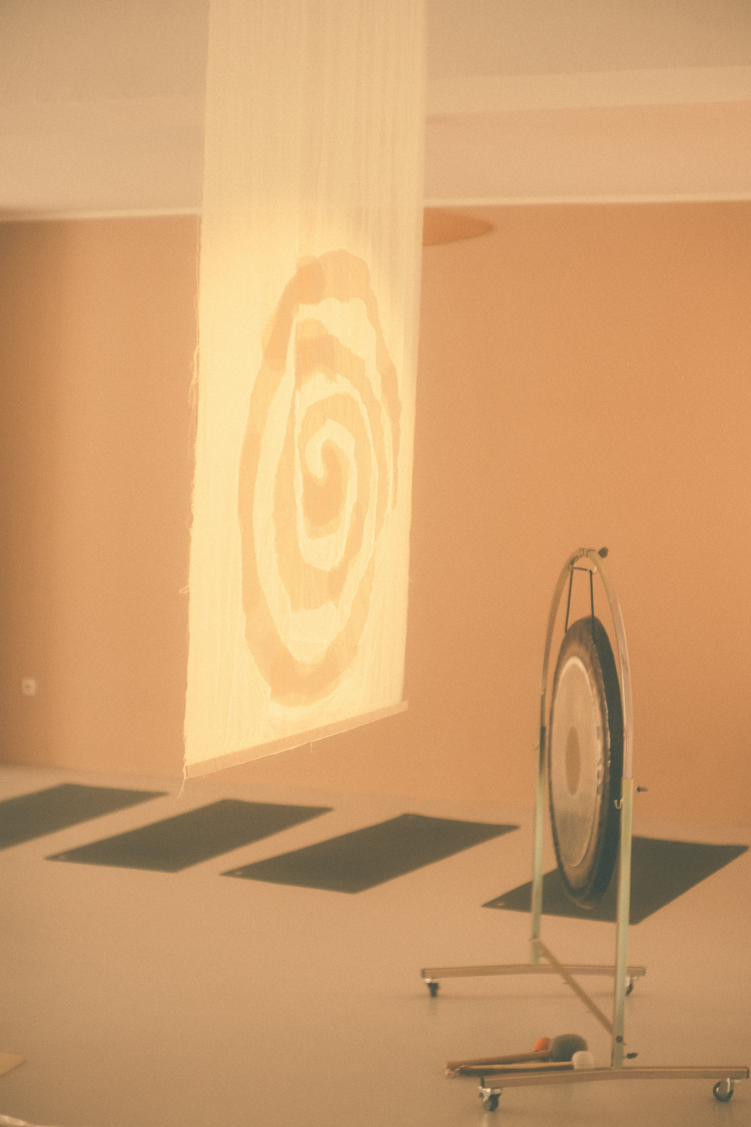 A gong on a stand in a room with beige walls. The room has a shadow of a spiral pattern projected on the wall.