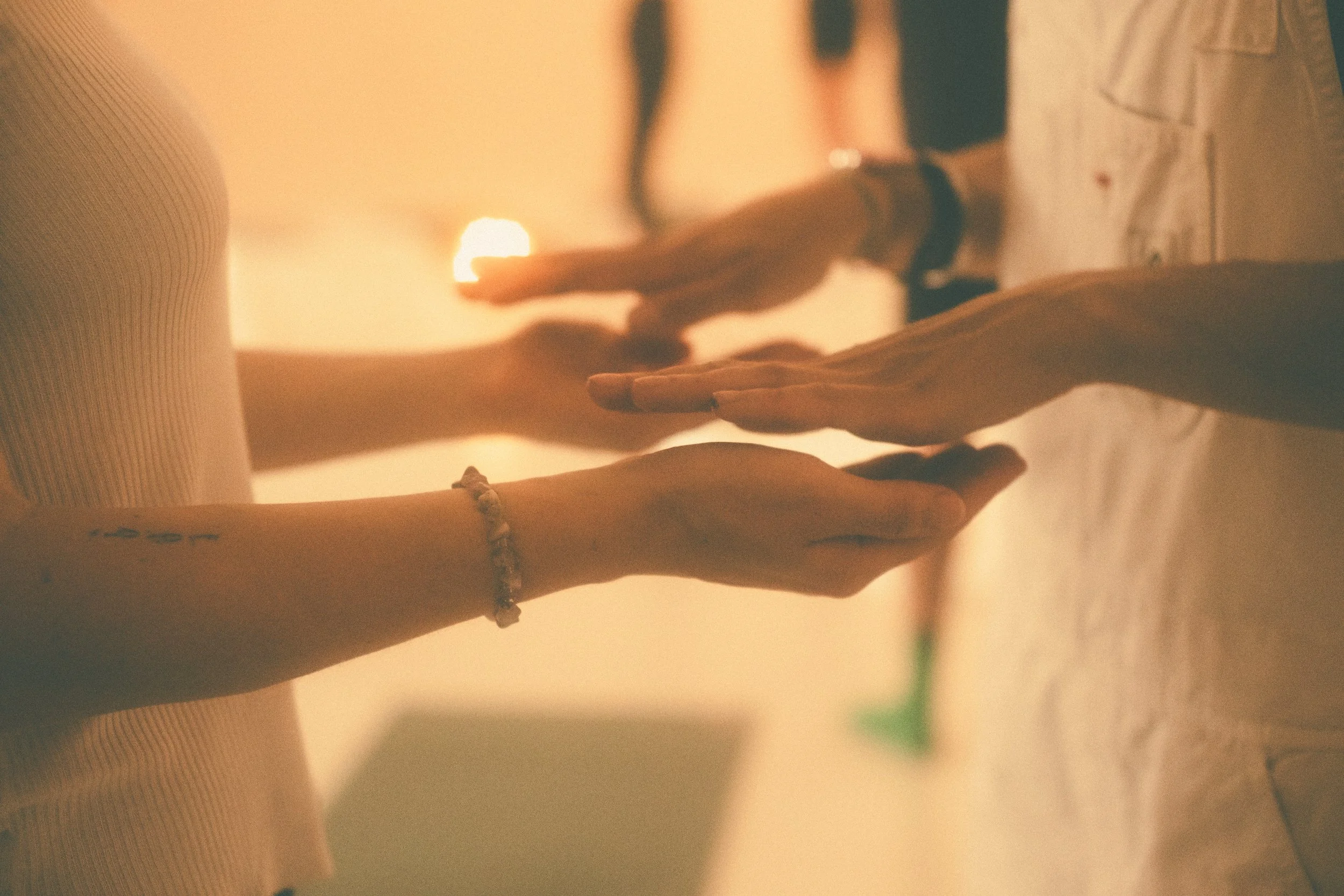 Two people practicing partner yoga on a bed with sunset light in the background, showing their hands and arms reaching towards each other.