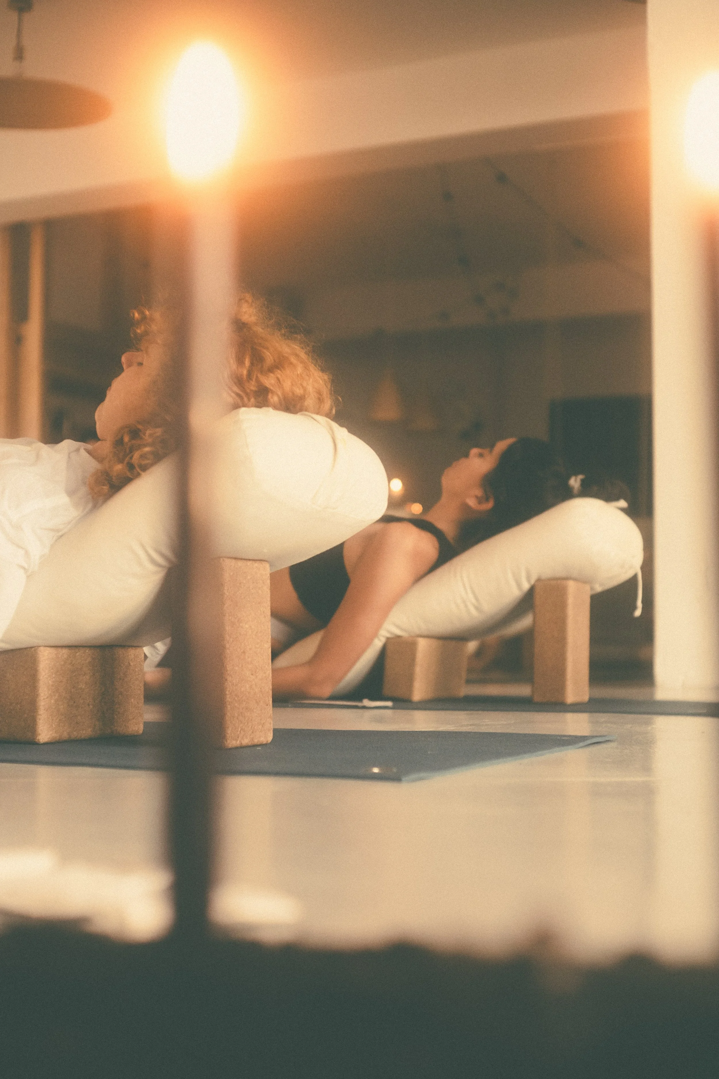 Two women practicing yoga on cushions with wooden supports, lying on their backs in a dimly lit room, with candles providing a warm glow.