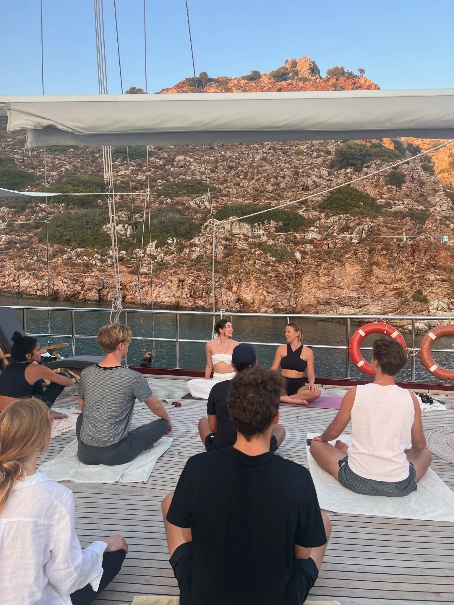 A group of people participating in a yoga or meditation session on a boat deck overlooking rocky hills and water at sunset.