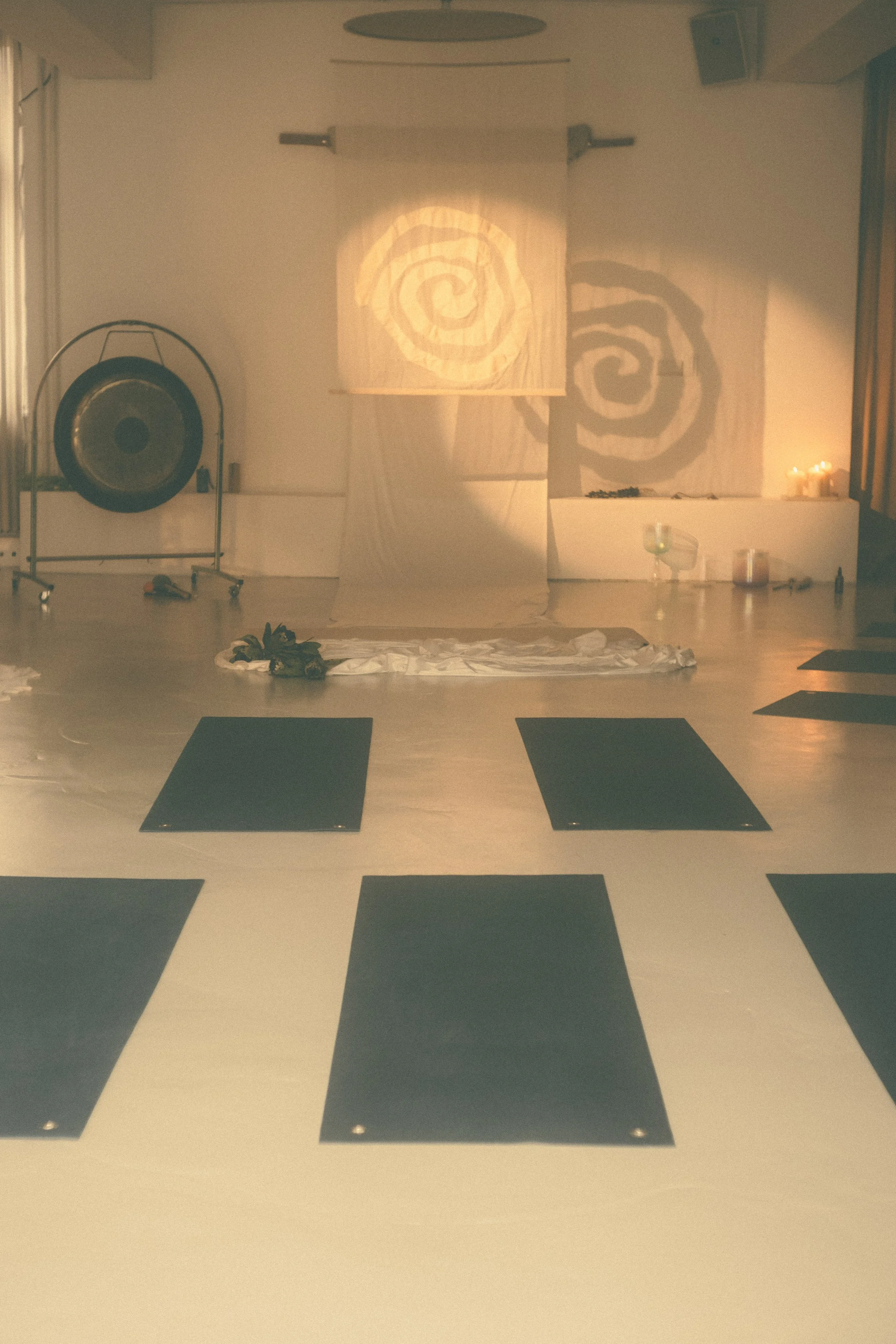 Yoga mats on the floor in a dimly lit room with candles, a gong, and a fabric backdrop with spiral patterns.