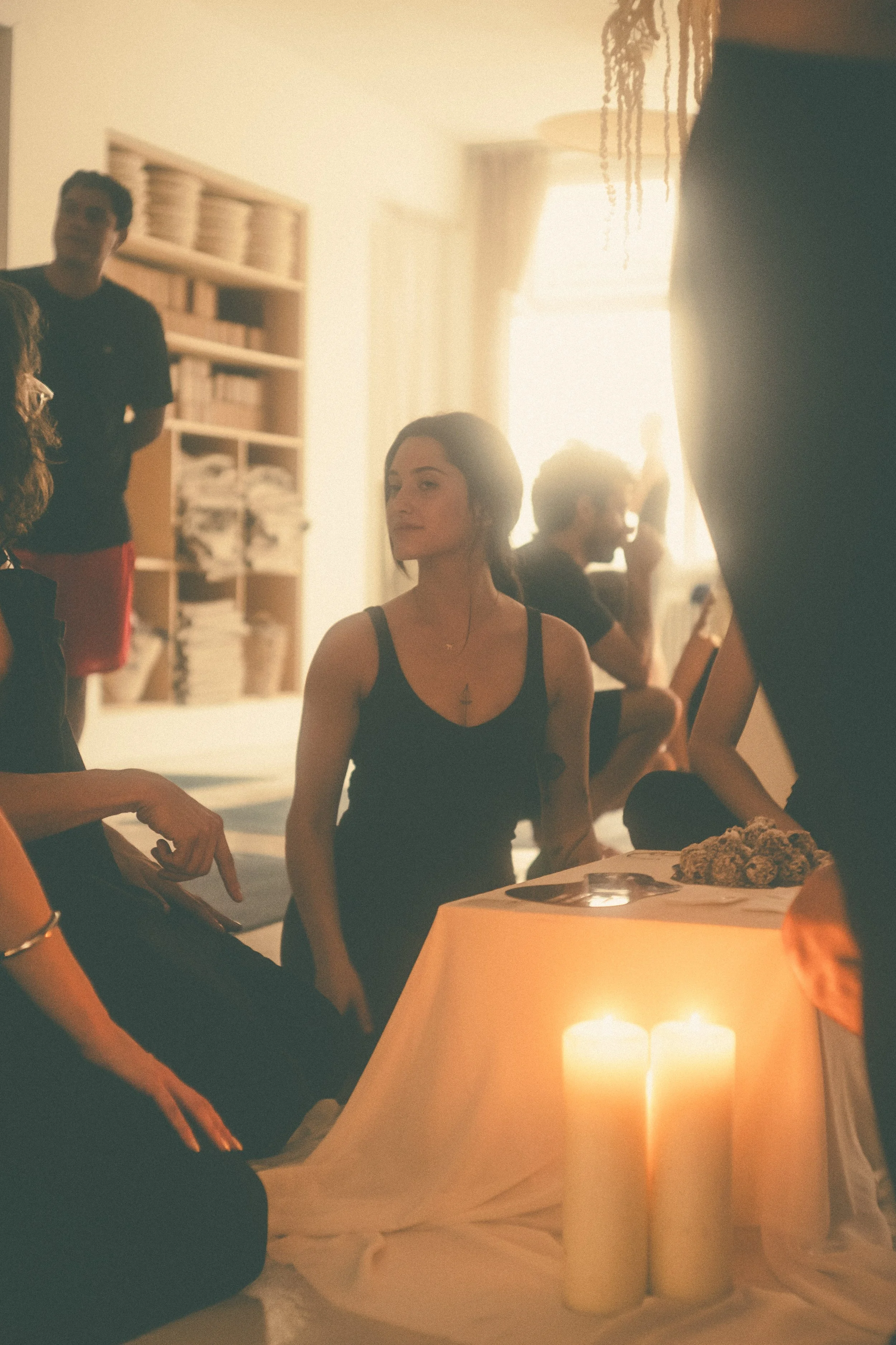 People gathered in a softly lit room, with candles on a table, some participating in a calming or meditative activity, and others observing thoughtfully.