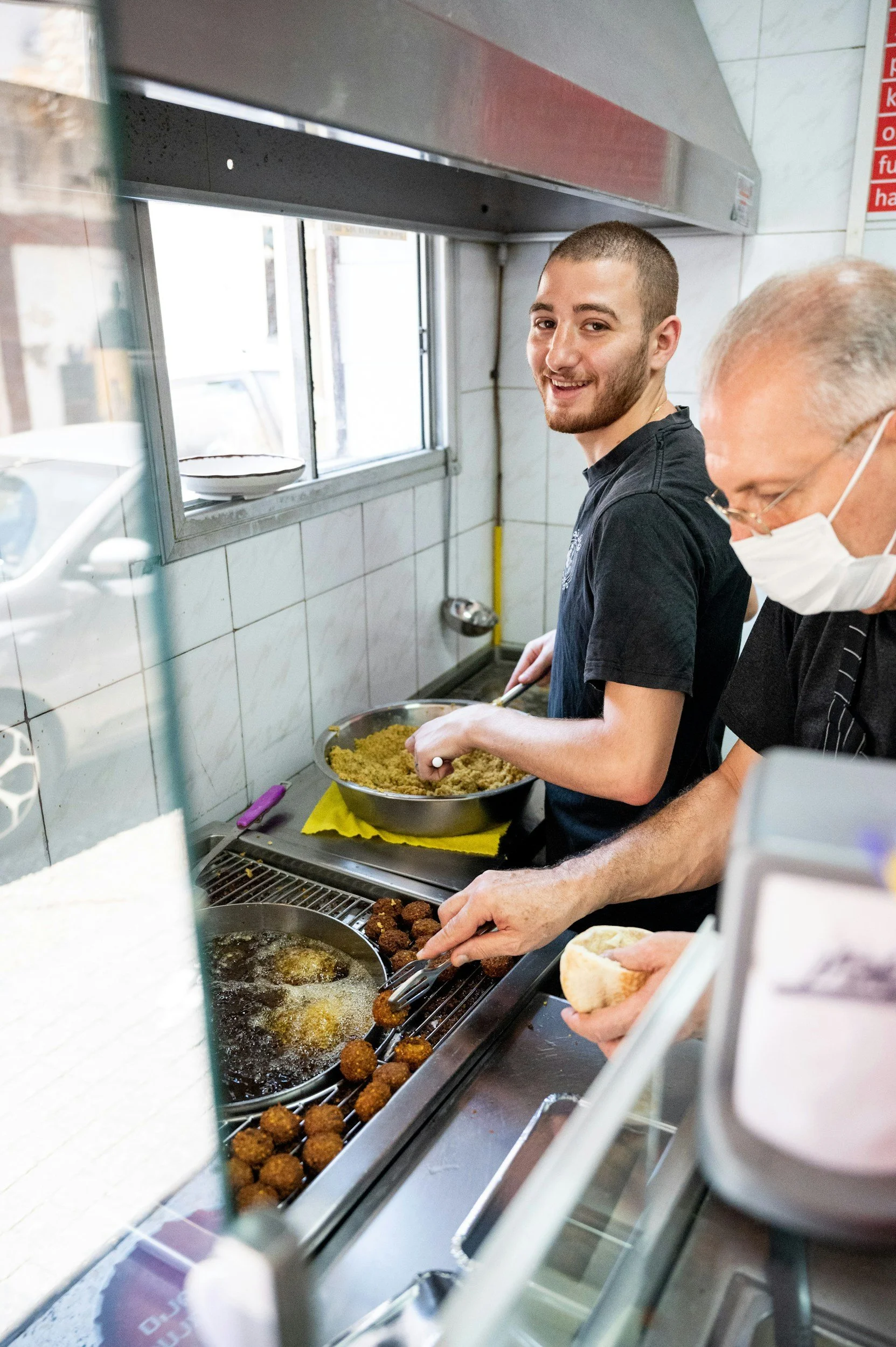Two men working in a kitchen, one smiling at the camera and the other wearing a face mask, preparing falafel and pita bread near a frying station.