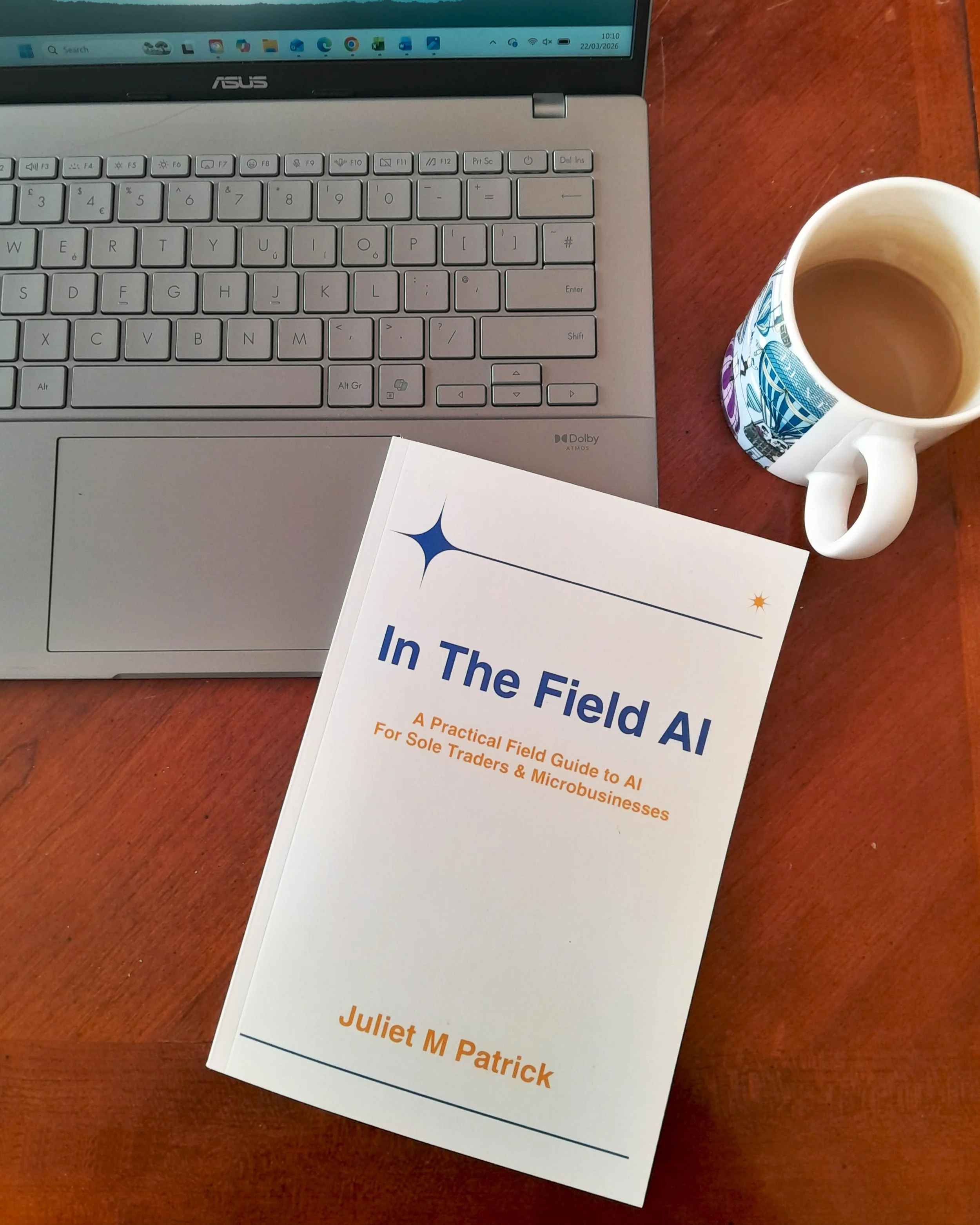Paperback book "In The Field AI" by Juliet M Patrick — a practical AI guide for sole traders and microbusinesses — on a desk with a laptop and cup of tea.