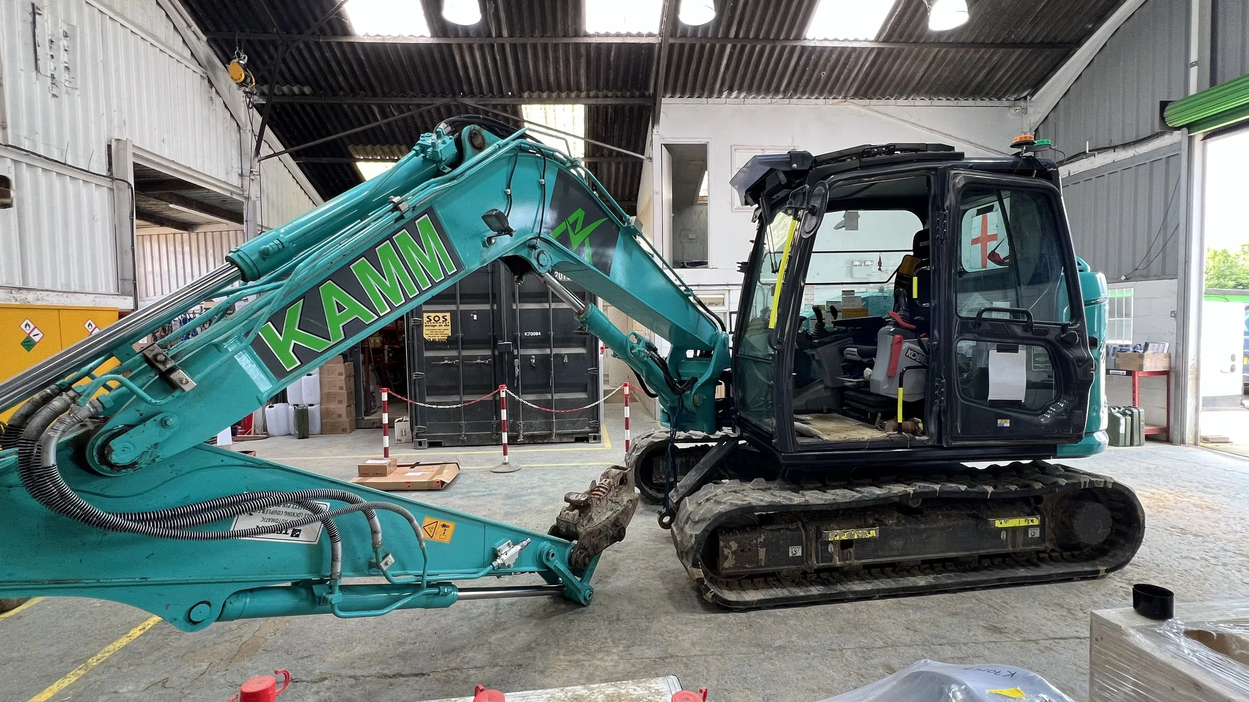 A teal KAMAZ mini excavator with a black cab, parked inside a warehouse, with various tools and equipment around.