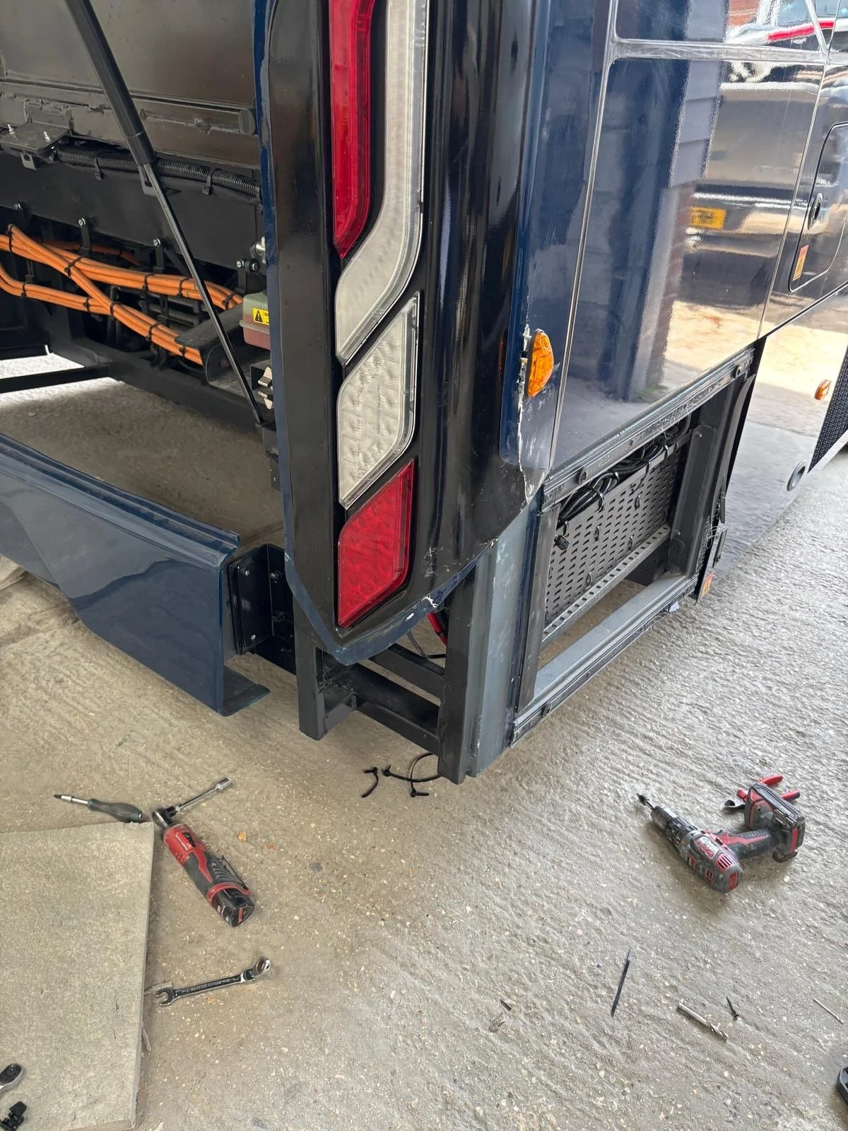 Close-up of the rear corner of a blue electric delivery vehicle, with work tools such as screwdrivers and a wrench on the ground nearby, and wires visible inside the open compartment.