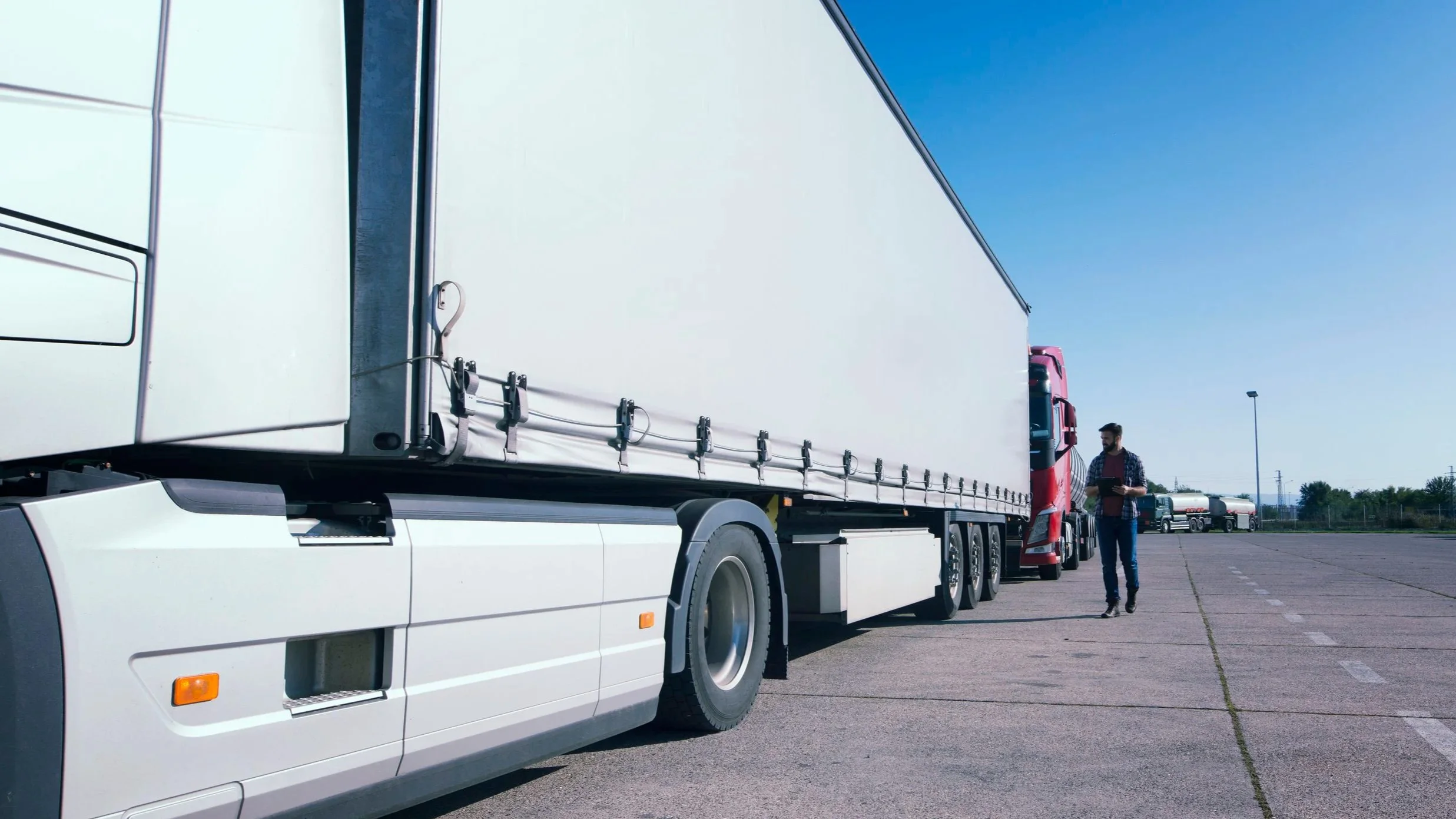 A person walking next to a large white semi-truck with a red cab, parked in a lot under a blue sky.