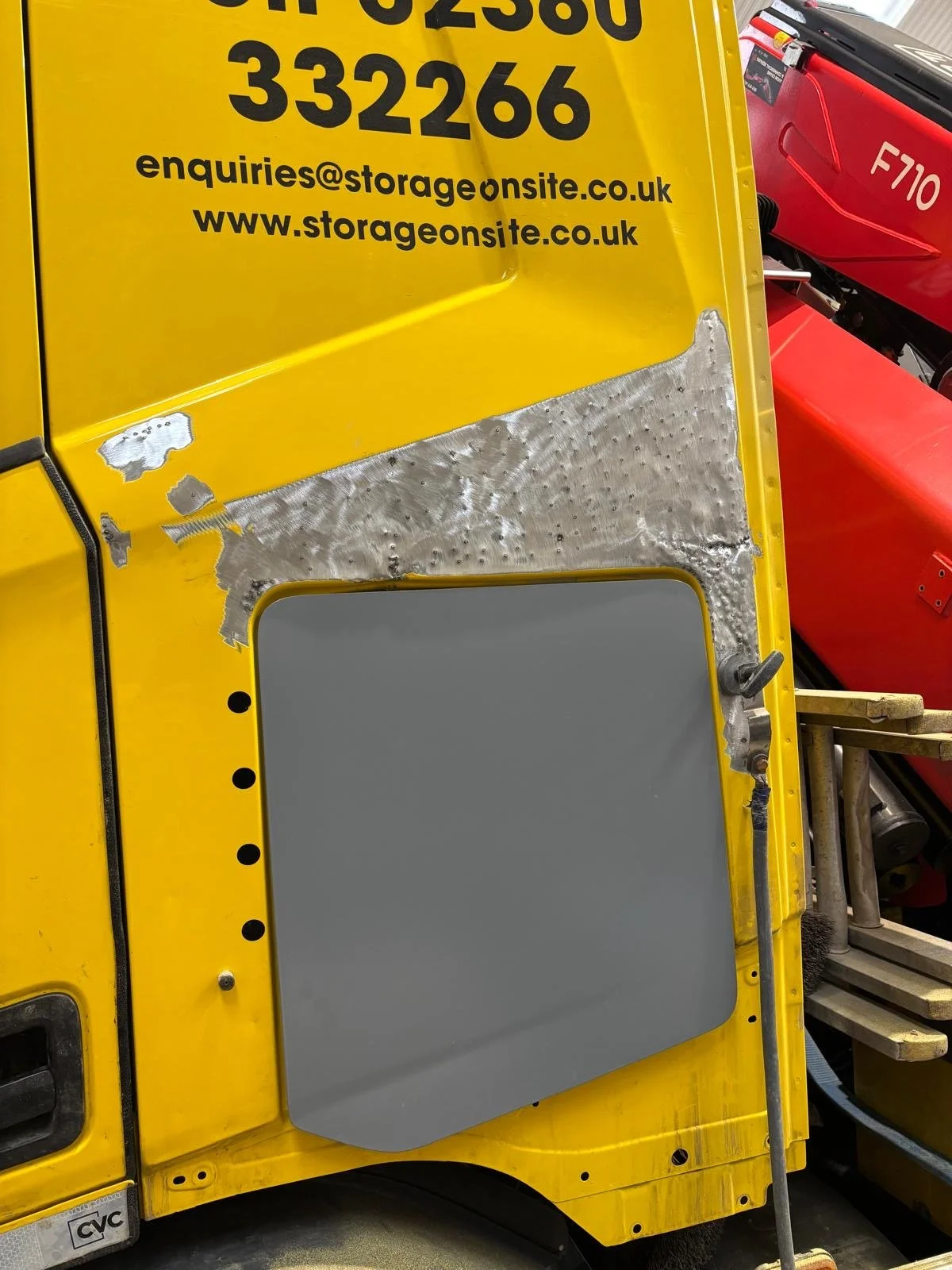 Close-up of a yellow van with black text displaying contact information, showing a patch of repair or modification with metal and sanding marks, and part of a red vehicle in the background.