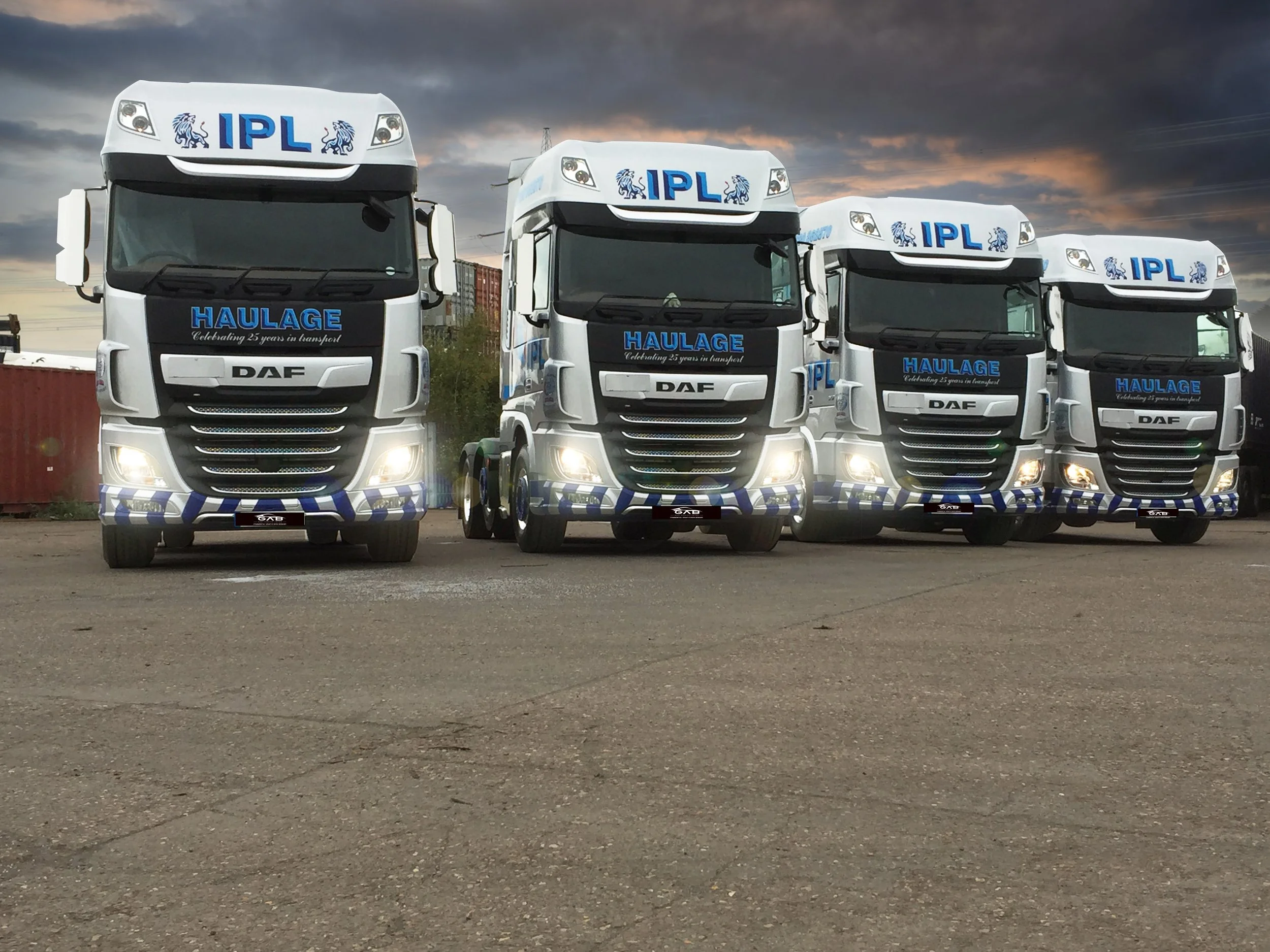 Four large trucks parked side by side on an asphalt lot at sunset. They are DAF trucks with white cabs, decorated with blue graphics and text, including
