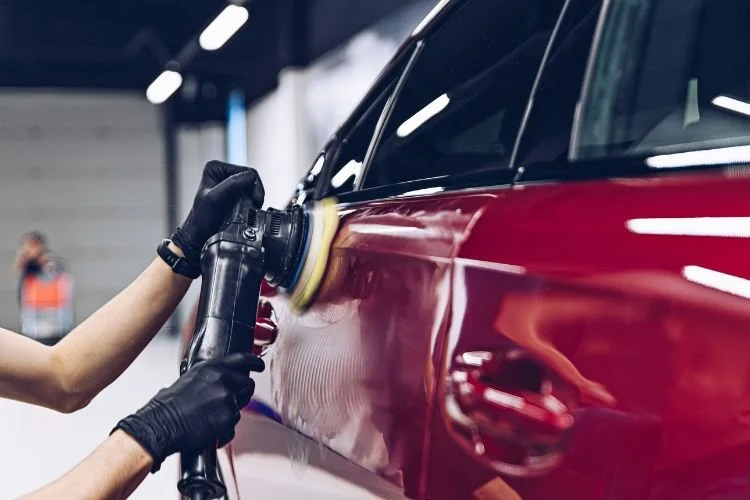 A person wearing black gloves polishing a red car with a circular buffer tool in an indoor auto detailing shop.