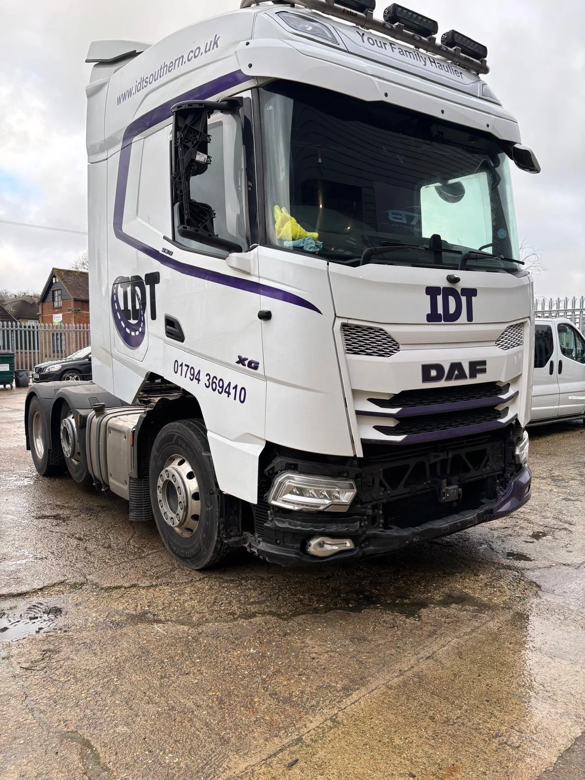 White DAF tractor truck with company logo and contact info parked on a wet, muddy ground, with some damage to the front bumper and grille area, and a small container on the trailer.