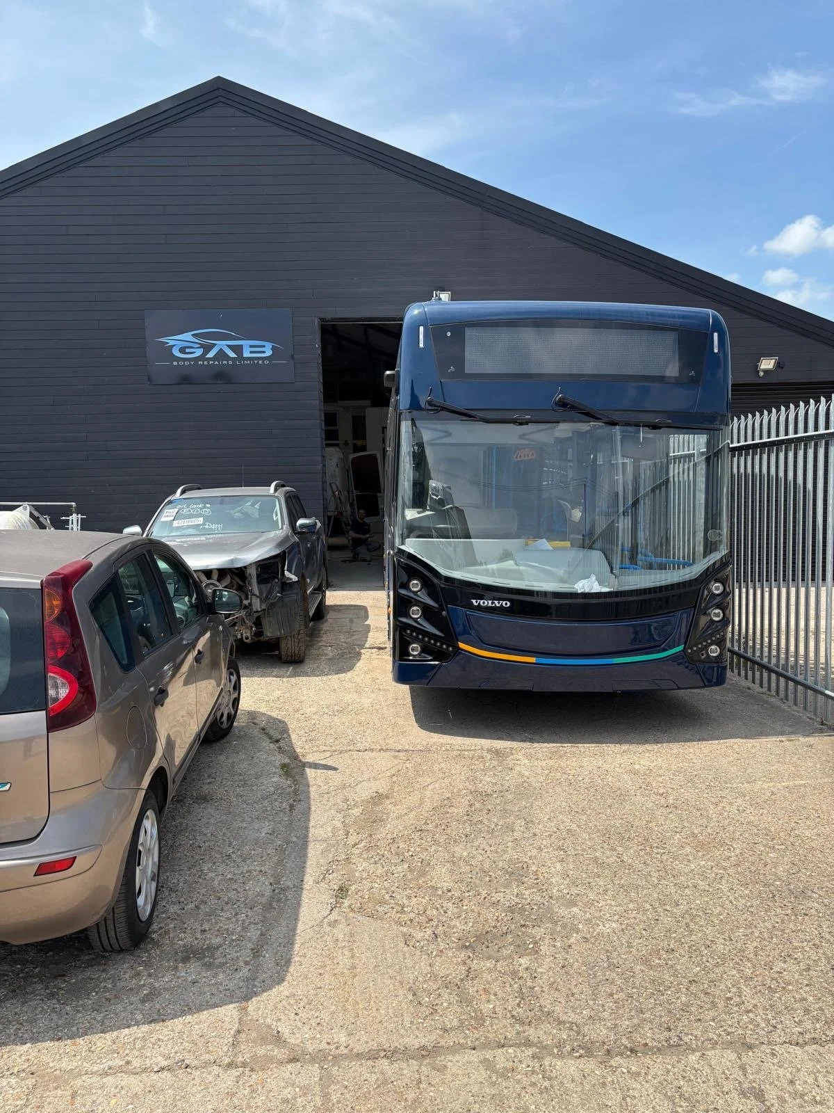 A blue Volvo bus parked outside a black body repair shop with a sign that reads 'GAB Body Repairs Limited.' To the left of the bus, there are two damaged cars, one silver and one beige, parked on a rough concrete surface. The background features a clear blue sky.
