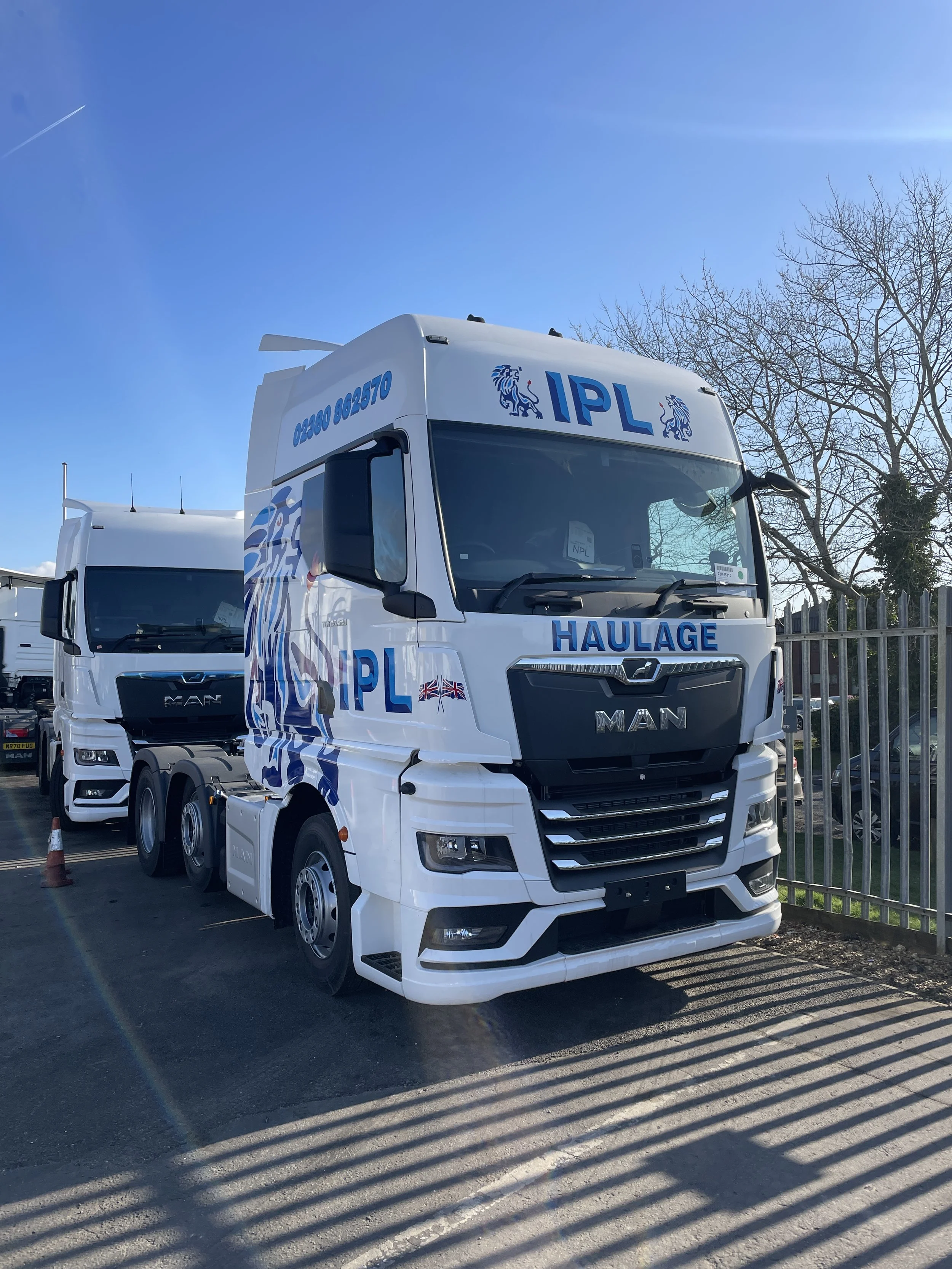 White truck with blue and black lettering and graphics, large grille, parked outdoors on a sunny day, with a metal fence on the right and other trucks behind it.