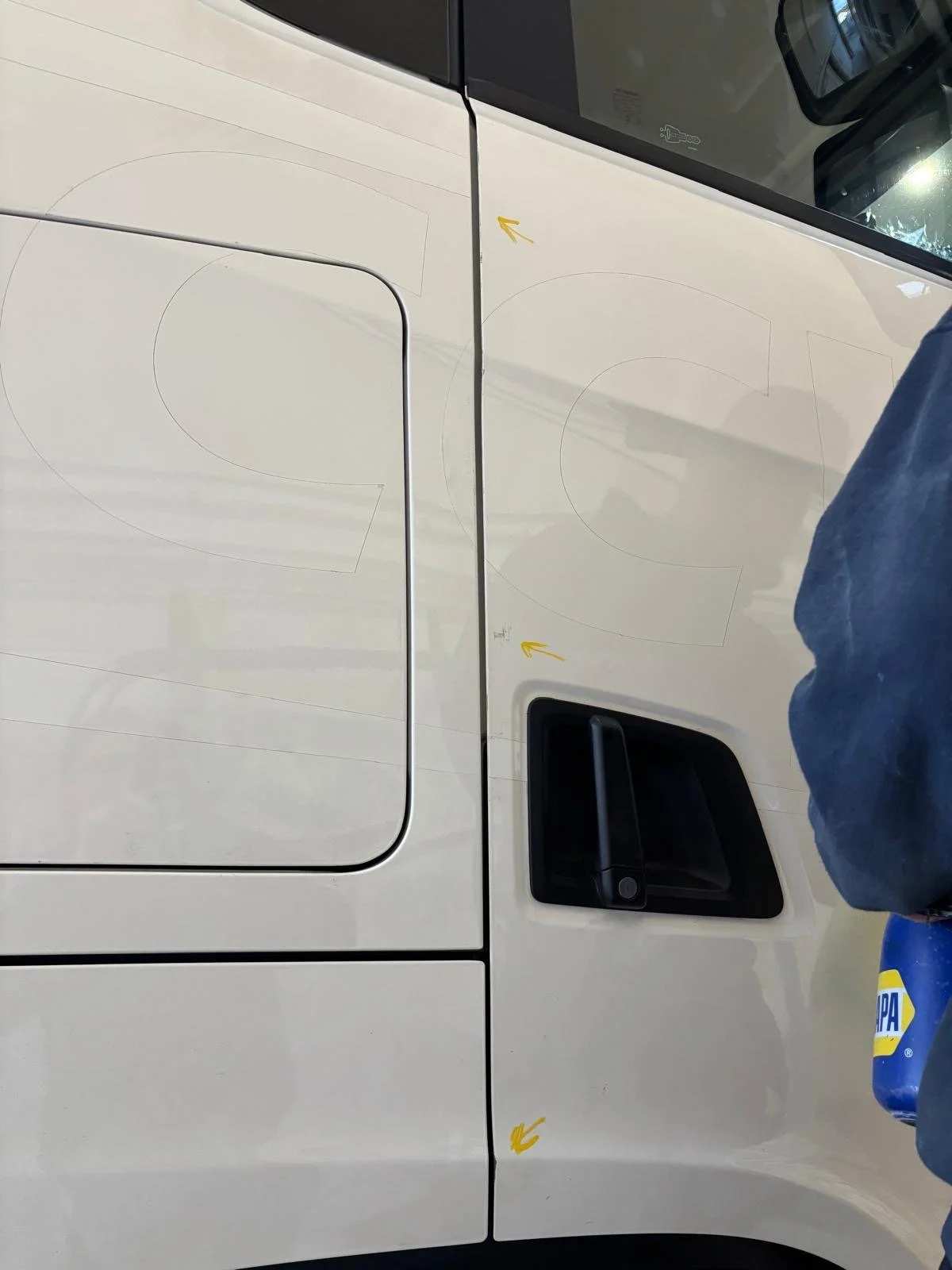 Close-up of a white vehicle's side door with yellow markings and a black handle, with a person wearing a dark jacket and holding a blue container nearby.