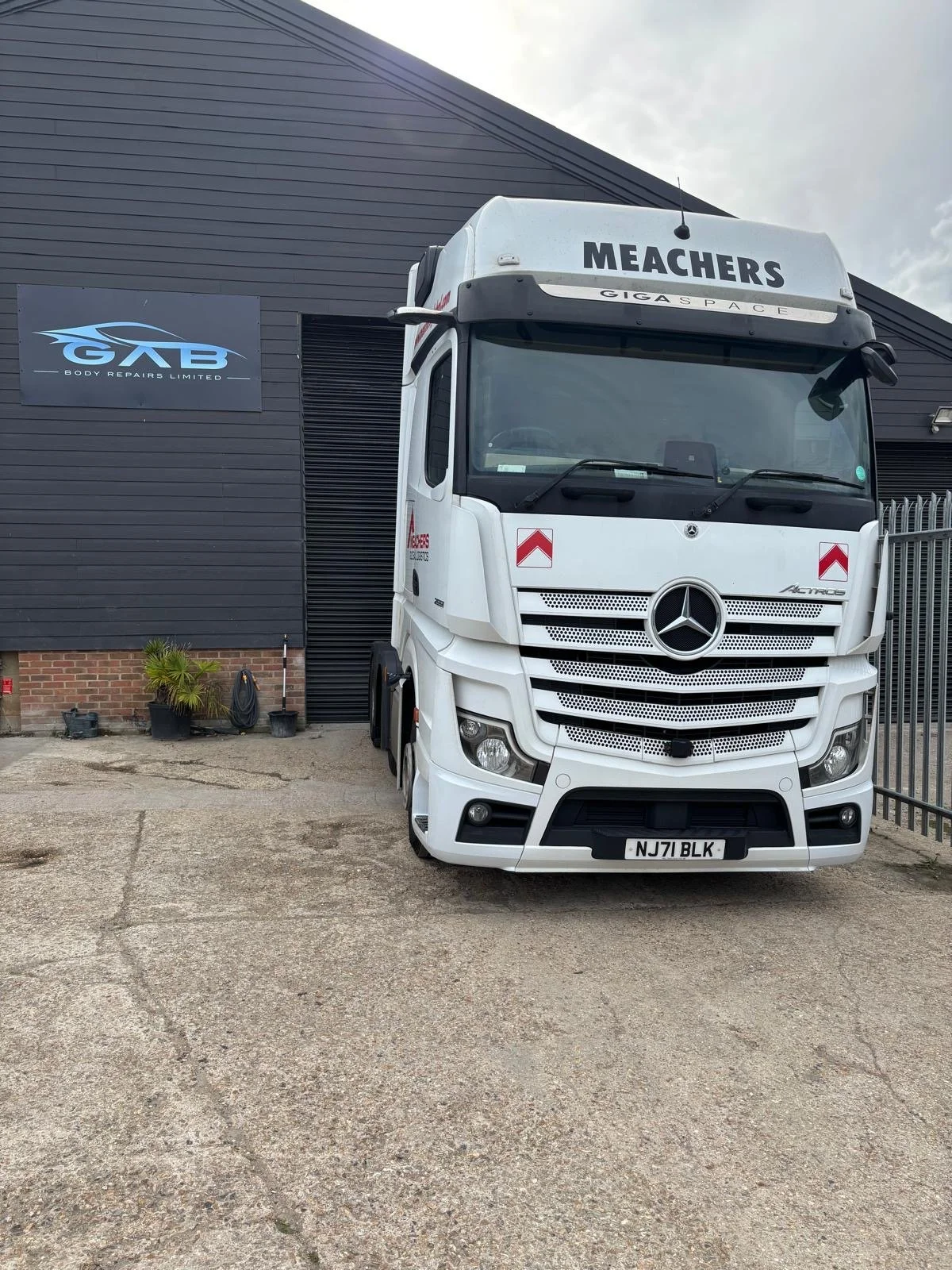 White Mercedes-Benz truck parked outside a building with black siding and a sign that reads Body Repairs Limited.' The truck has the word 'MEACHERS' on the top and red reflective tape on the front. The ground is a concrete surface with some cracks an