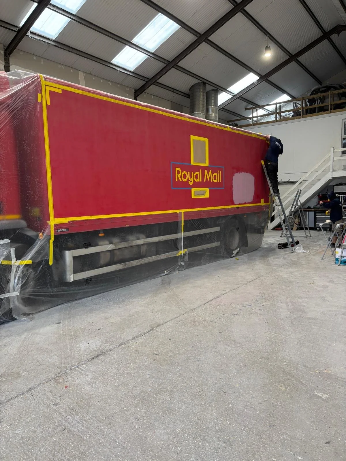 A large red Royal Mail truck is being painted inside a workshop. A person is standing on a ladder working on the upper part of the truck, with plastic covering around the vehicle to protect it during painting.