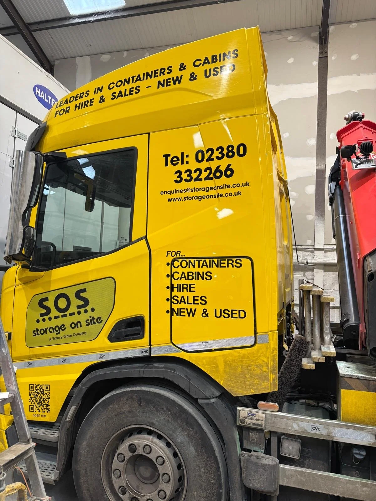 Yellow truck with signs advertising storage containers and cabins for hire and sale, including new and used options. Contact details and a QR code are displayed on the side of the truck, which appears to be inside a warehouse or storage facility.