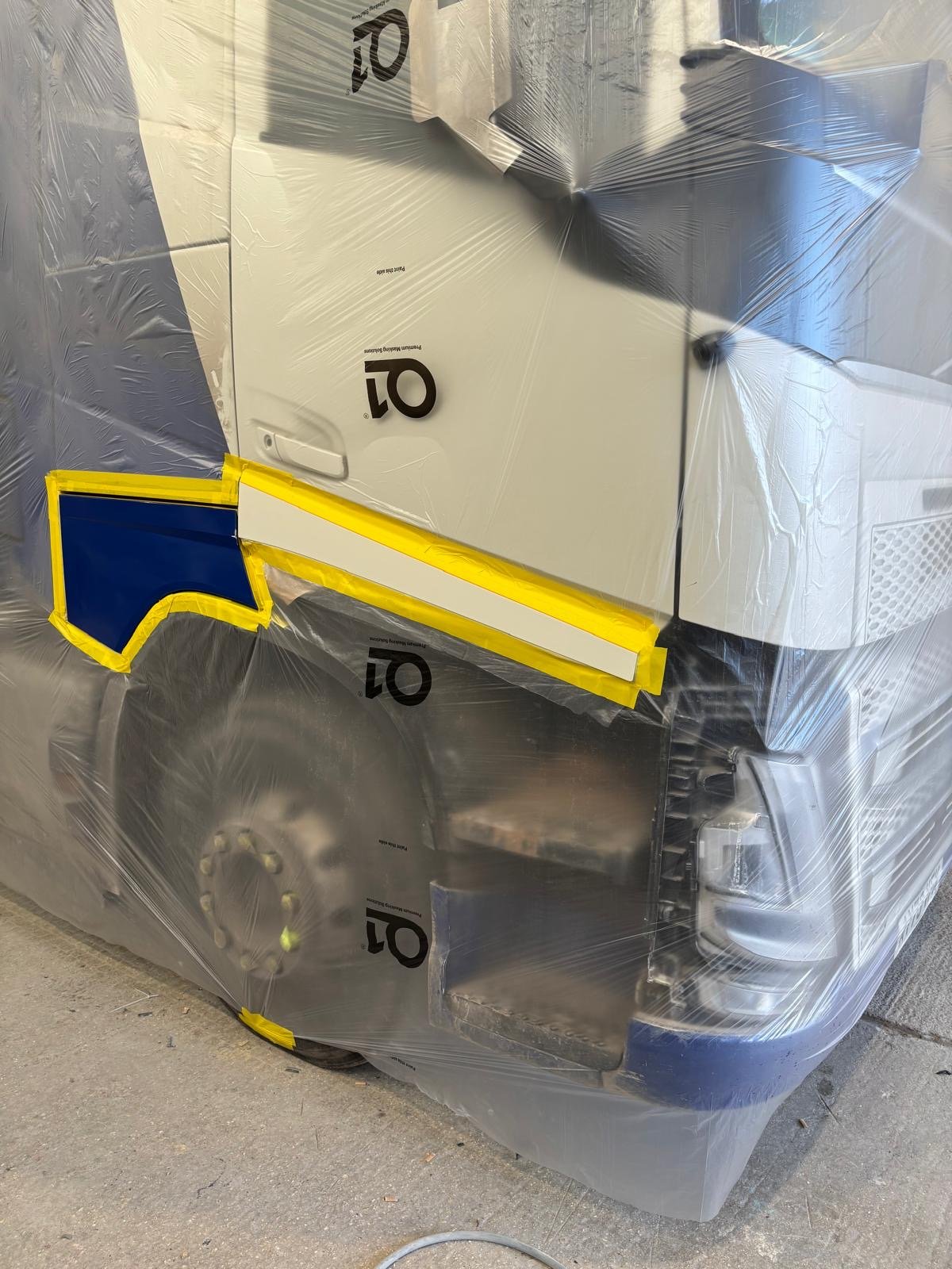 Front of a large vehicle, possibly a truck or bus, wrapped in clear protective plastic with part of a blue and yellow logo visible on the side.