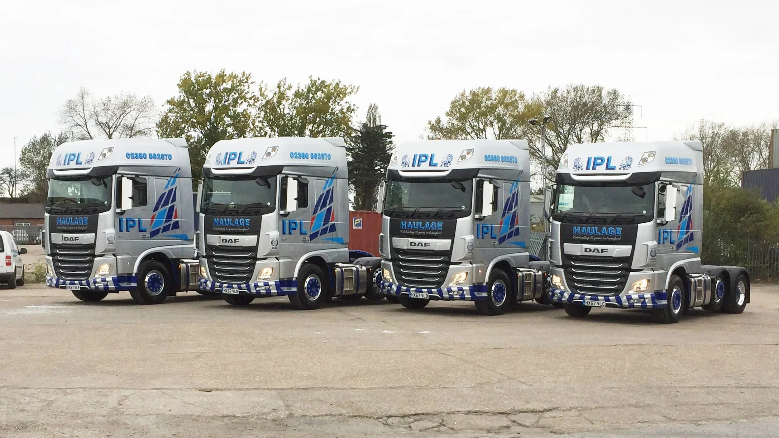 Four identical silver semi-trucks with blue and red graphics parked in a lot with trees and a cloudy sky in the background.