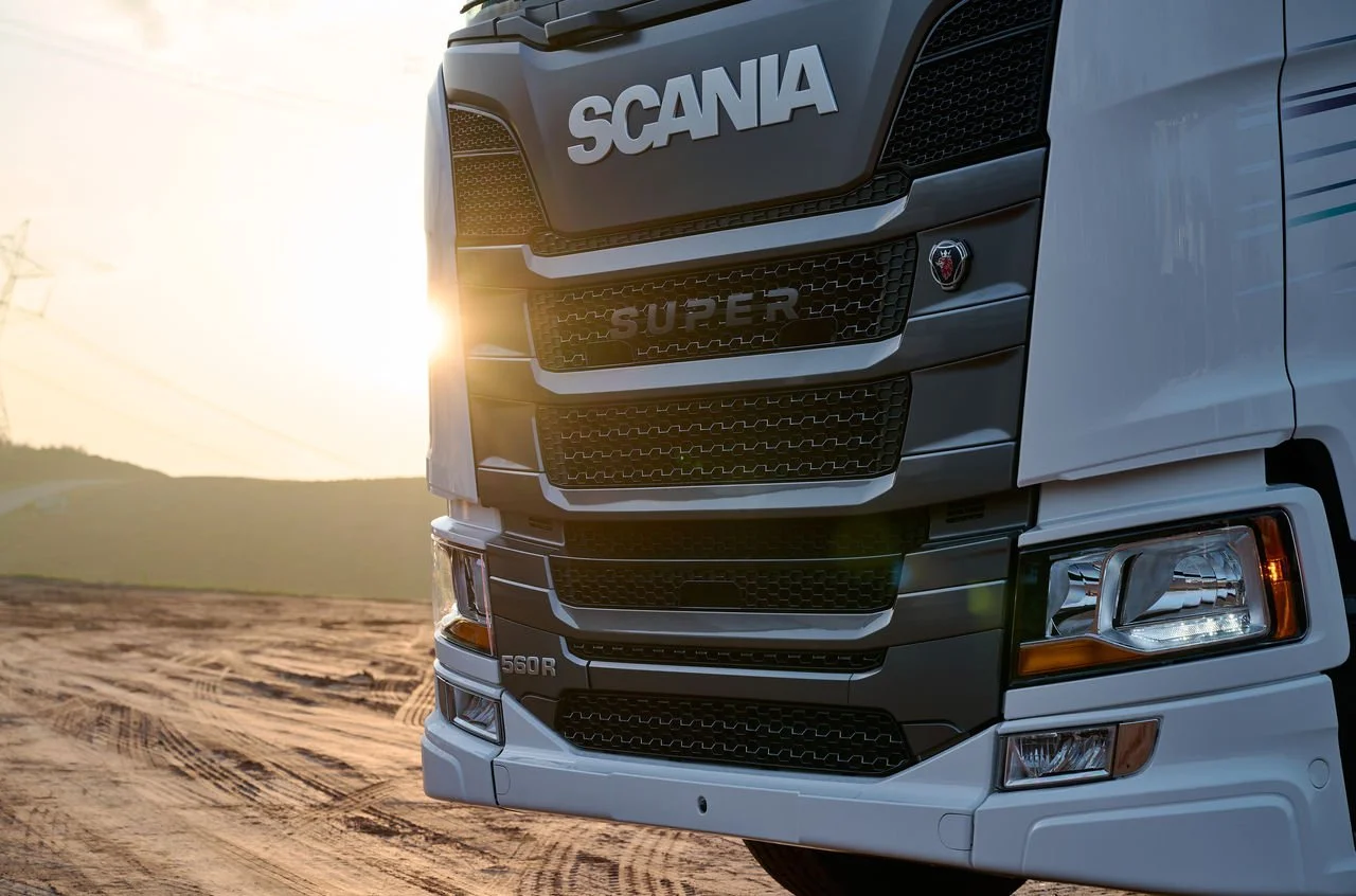 Close-up of the front of a white Scania semi-truck on a dirt road during sunset with hills in the background.