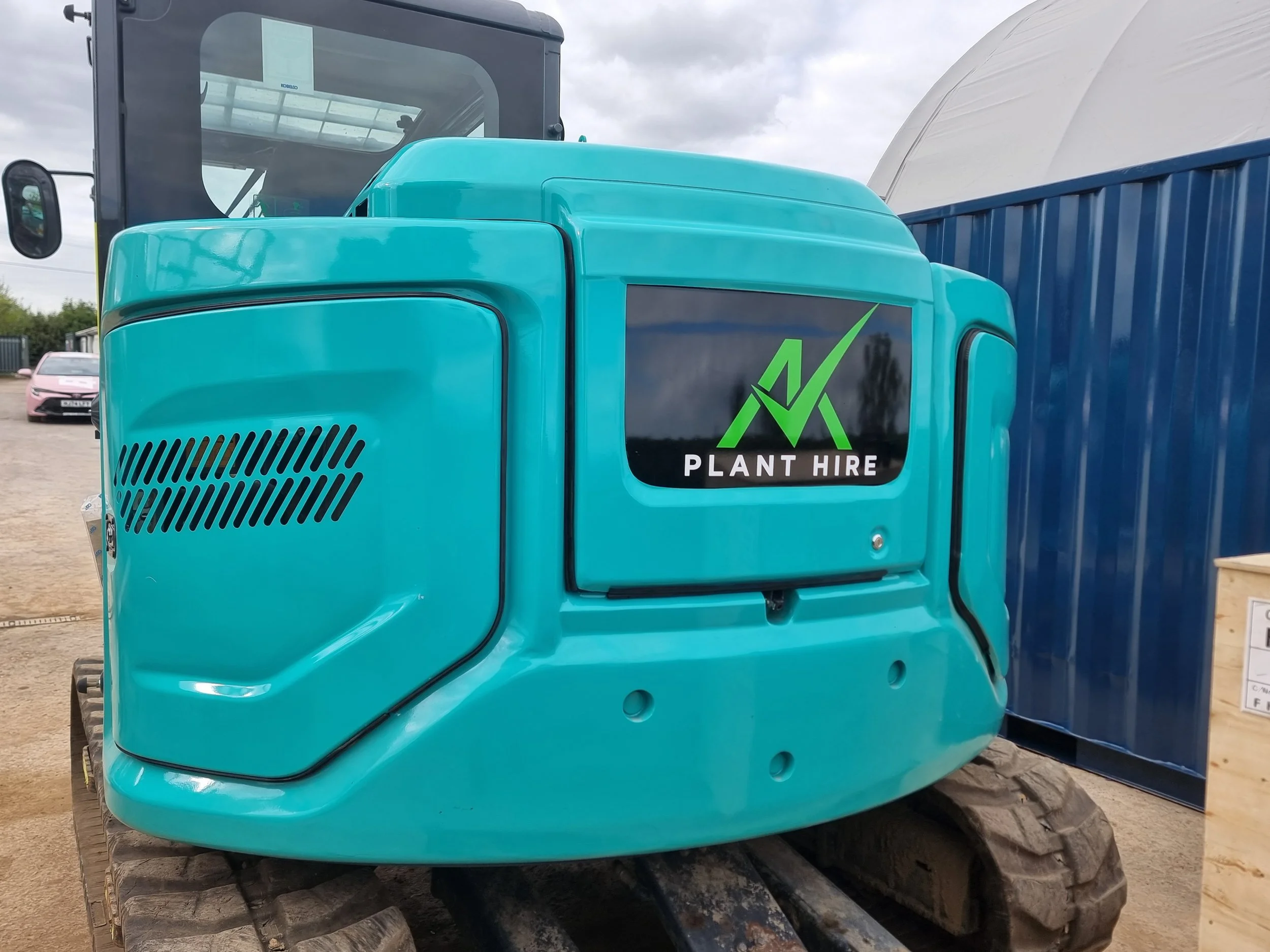 Construction mini excavator with a teal body, displaying a logo and the words 'PLANT HIRE' on the side, parked outdoors near a blue shipping container and other vehicles in the background.