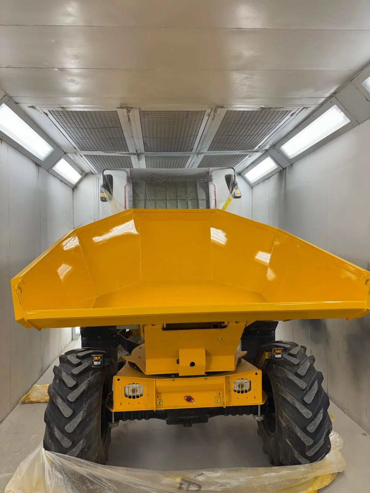 Yellow construction vehicle inside a spray booth, possibly a grader or similar equipment, with lighting on the ceiling.