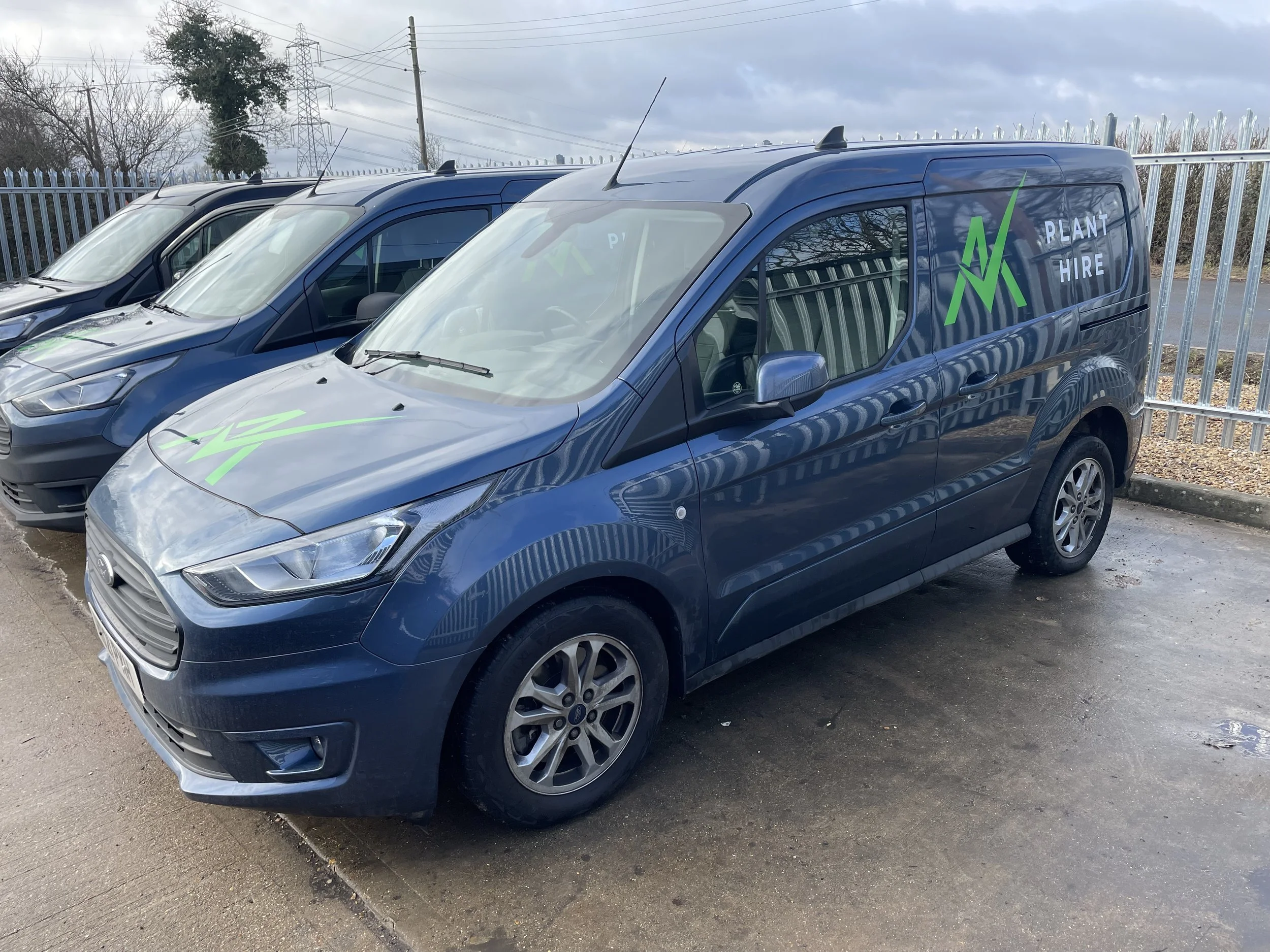 Blue commercial van with green and white logo and text reading 'Plant Hire,' parked in a lot beside other vehicles.