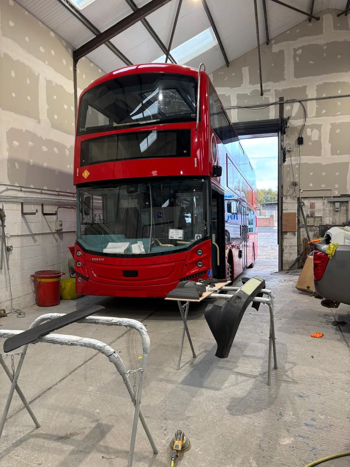 Inside a garage with unfinished walls, a red double-decker bus is parked. The bus is undergoing maintenance or restoration, with parts and tools around it.