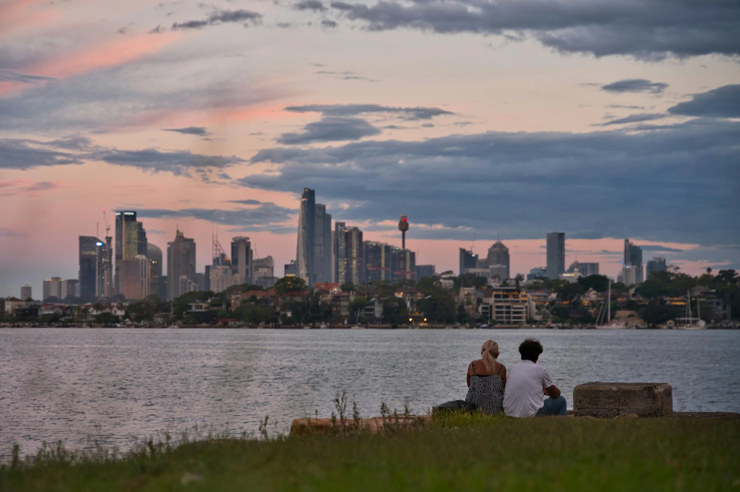 A couple sitting on the grass by a body of water, overlooking a city skyline at dusk with pink and gray clouds with tall buildings in the background.