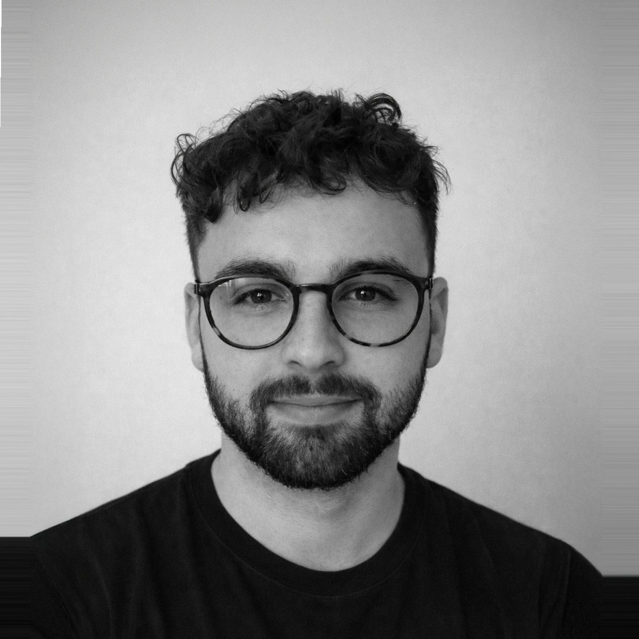 Black and white photo of a man with curly hair, glasses, and a beard, smiling slightly, wearing a dark t-shirt, standing against a plain background.