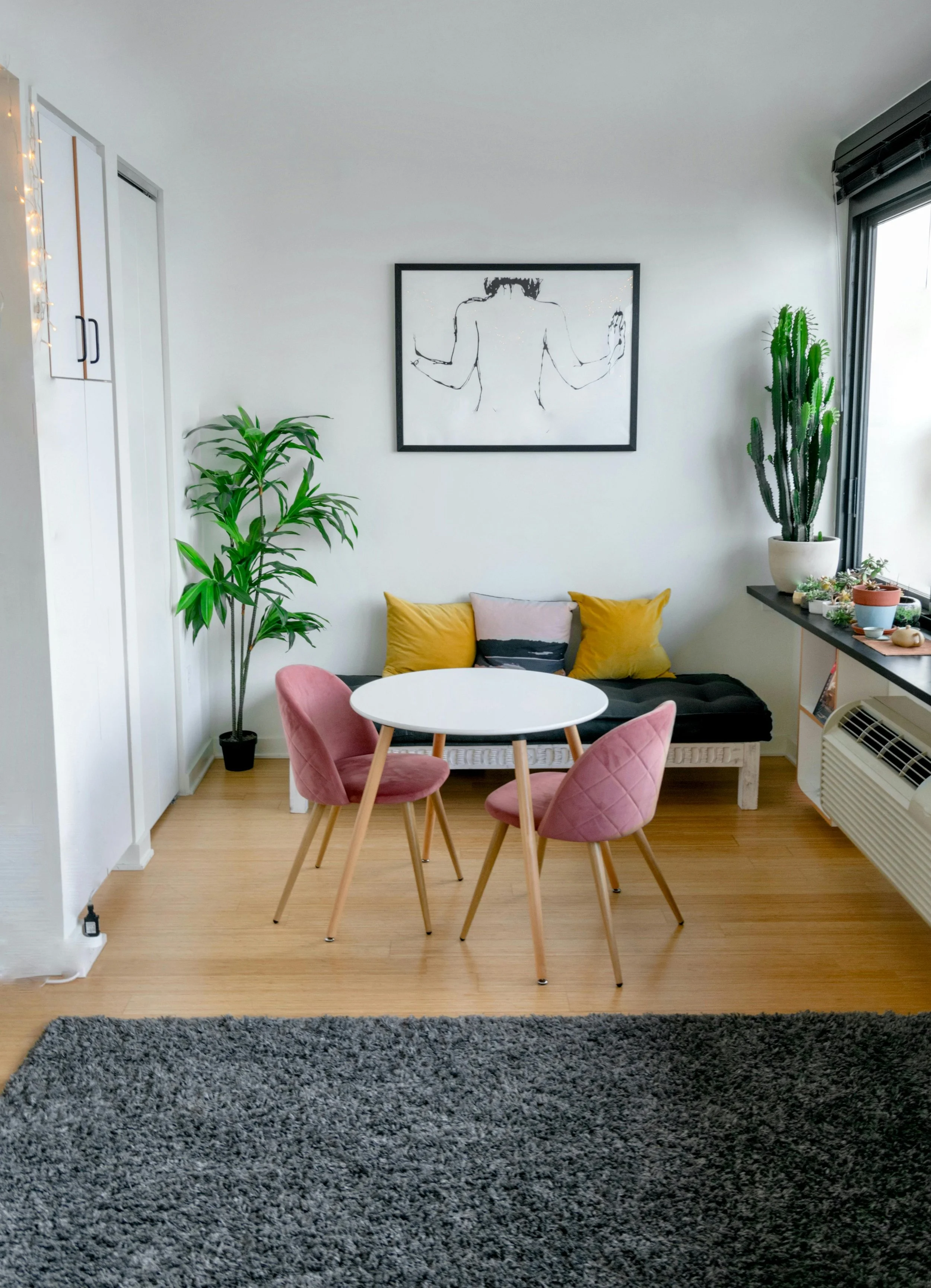 Cozy living room corner with a small round white table surrounded by three pink upholstered chairs, a black daybed with yellow pillows, a large potted cactus next to a window, and plants on the window sill, with a framed minimalistic line drawing of a person on the white wall.