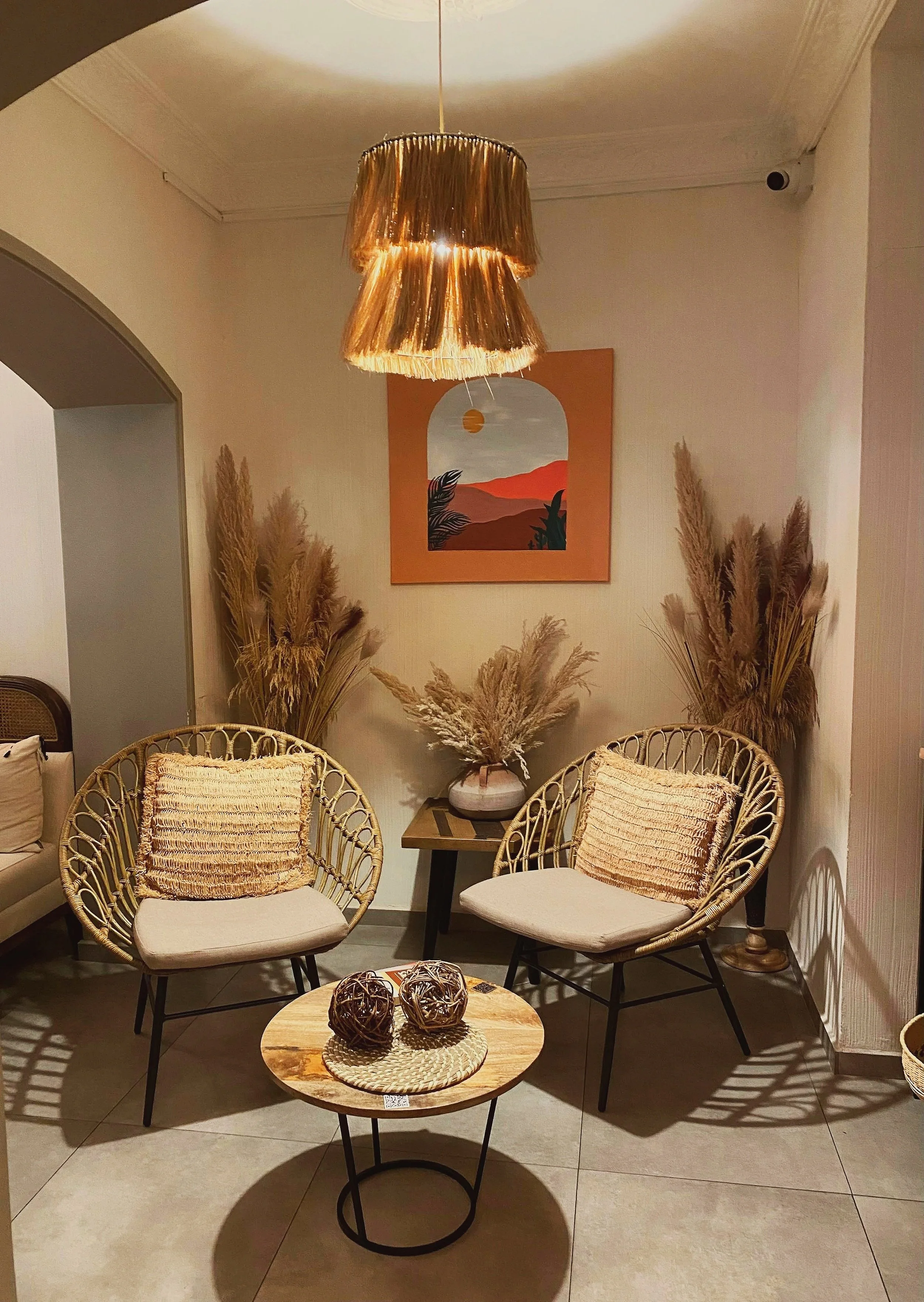 Living room corner with two rattan chairs with beige cushions and textured pillows, a small round wooden coffee table with decorative balls, a dark wood side table with pampas grass in a white vase, and a colorful landscape painting on the wall with warm earthy tones and minimalist decor.