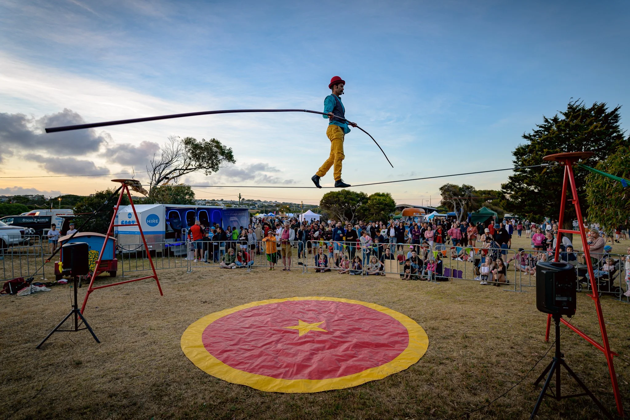 A circus performer walking on a tightrope in an outdoor fairground at sunset with a crowd watching behind a safety barrier.