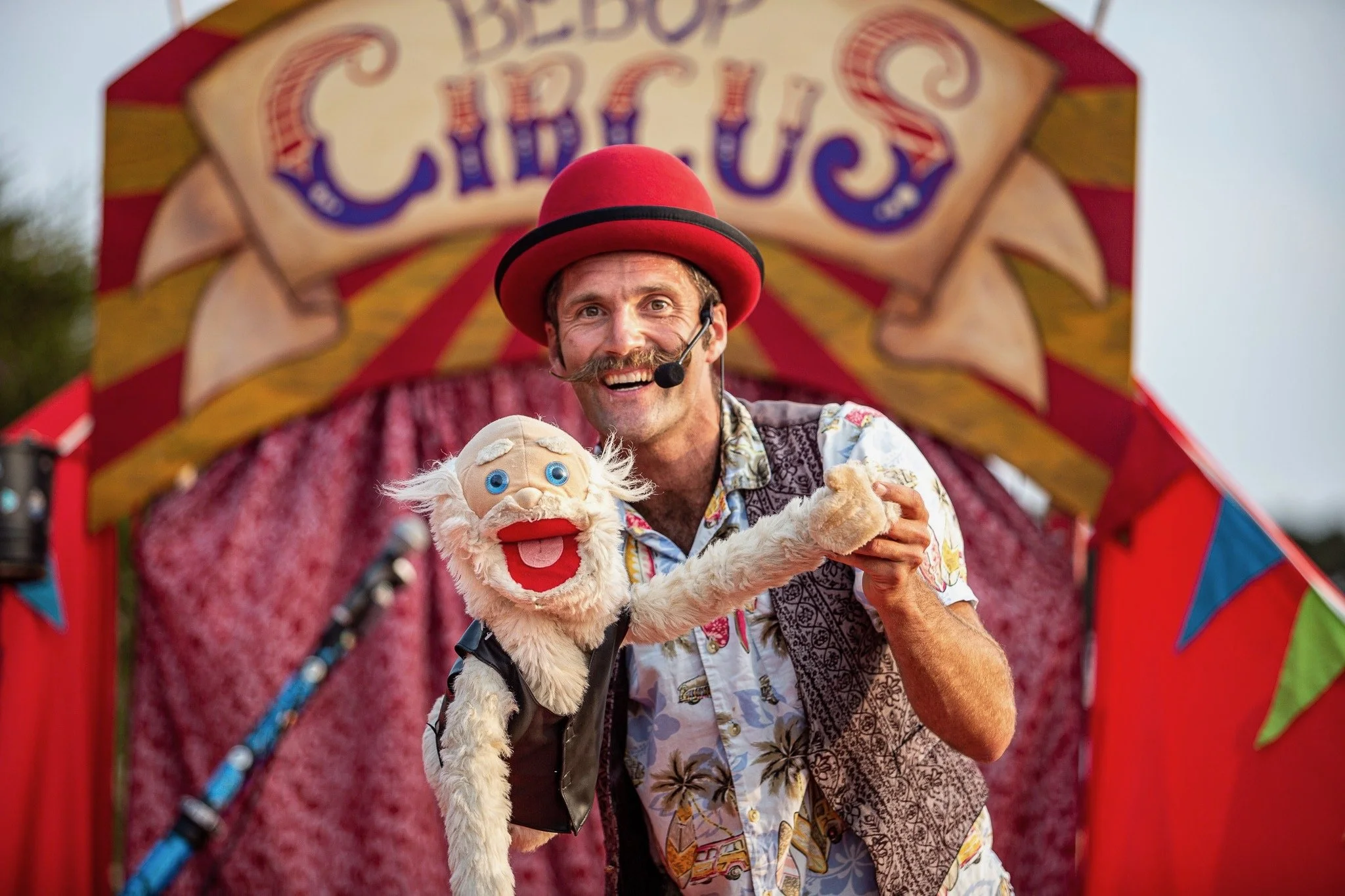 A man dressed as a clown performing on stage with a puppet at a circus, under a colorful circus tent, smiling and wearing a red bowler hat, a microphone headset, and a floral shirt.