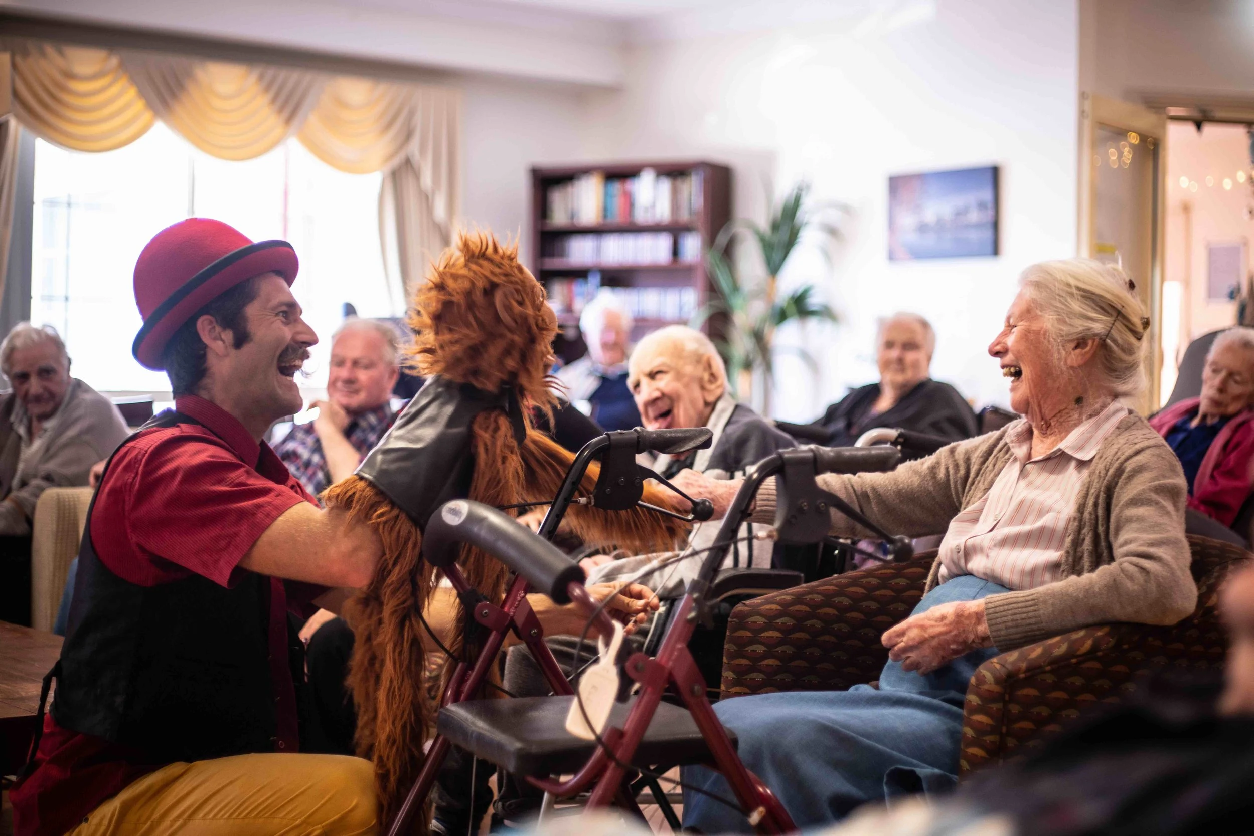 A man dressed as a clown with a red hat and yellow pants is holding an orange dog and entertaining elderly women in a nursing home. The women are smiling and laughing, enjoying the visit.
