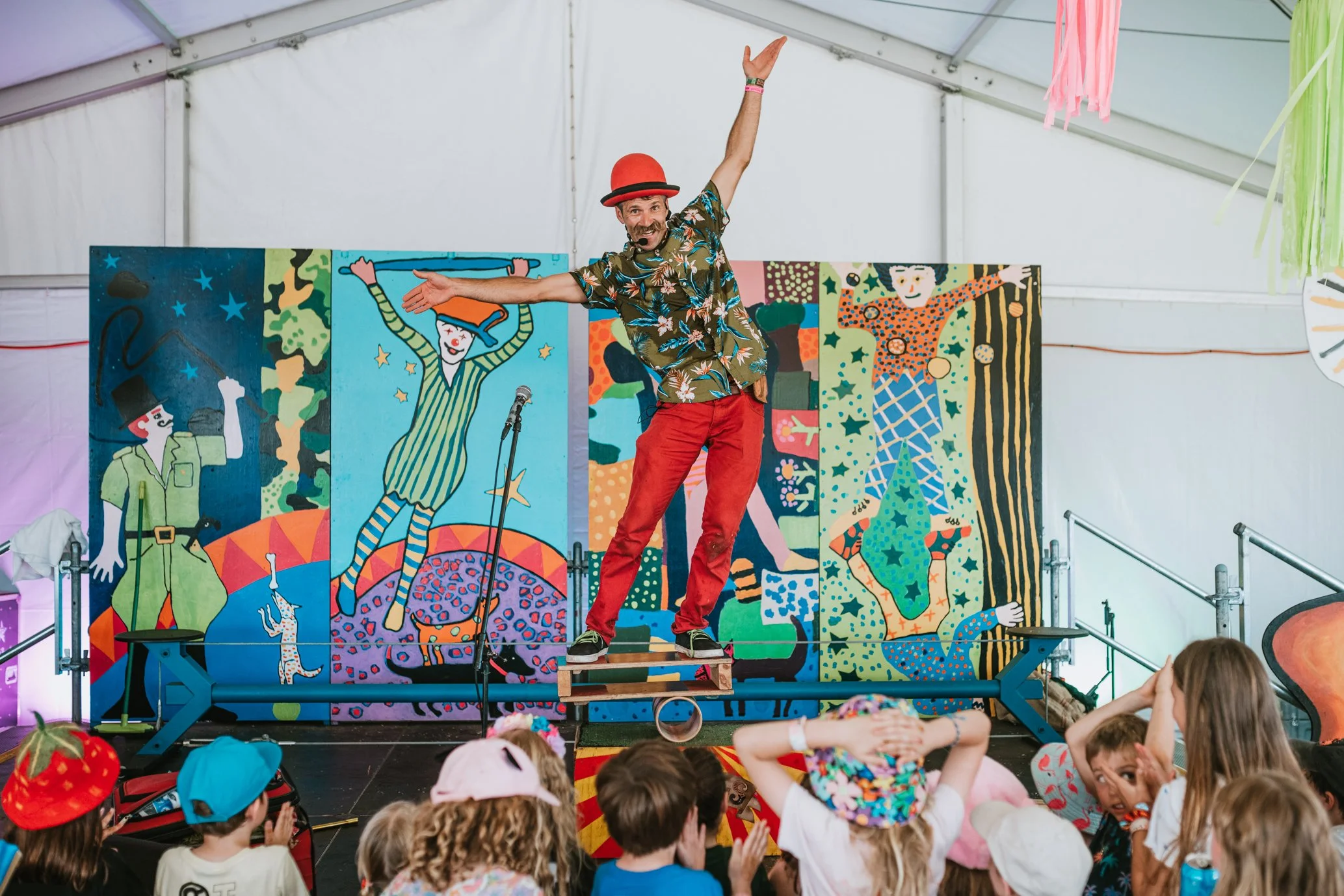 A clown in a red hat and colorful shirt performs on a small stage with children watching in front. The backdrop features vibrant, illustrated circus characters and animals.