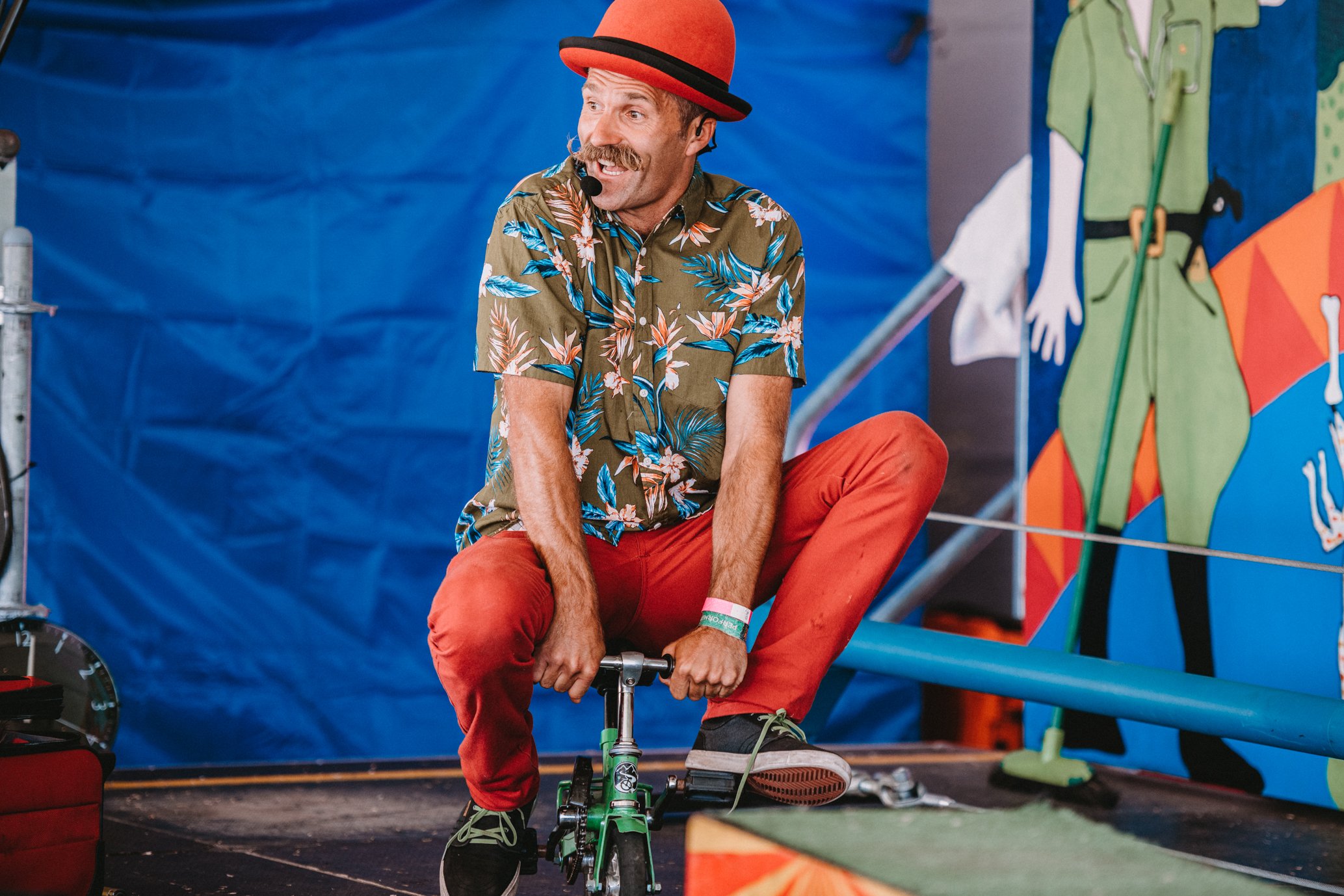 A performer dressed in a tropical shirt, red pants, and a red hat, sitting on a children's tricycle on stage during a circus act.