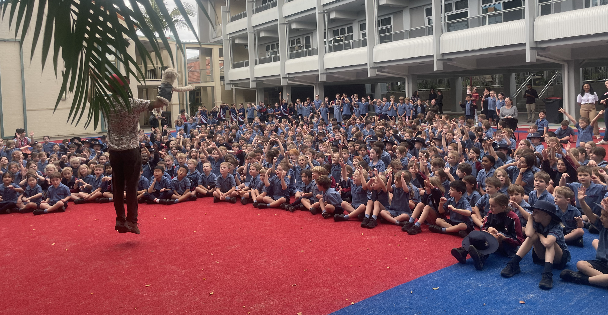 A large group of school children in uniforms sitting on a red and blue outdoor carpet, raising their hands, facing a performer in front of them, with a multi-story school building in the background.
