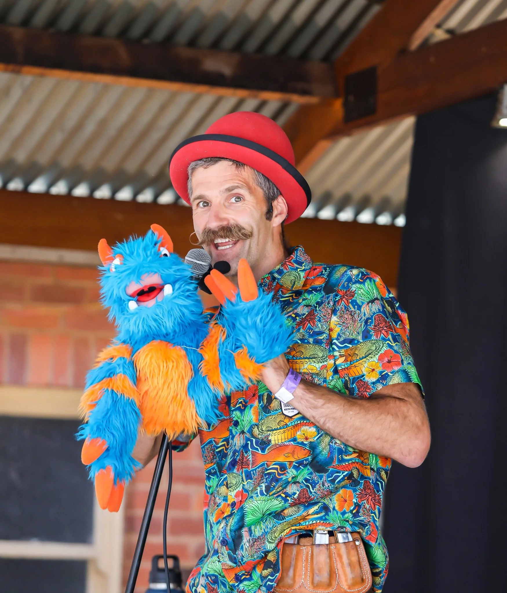 A man with a mustache wearing a colorful Hawaiian shirt, red hat, and holding a blue furry puppet with orange accents, speaking into a microphone indoors.
