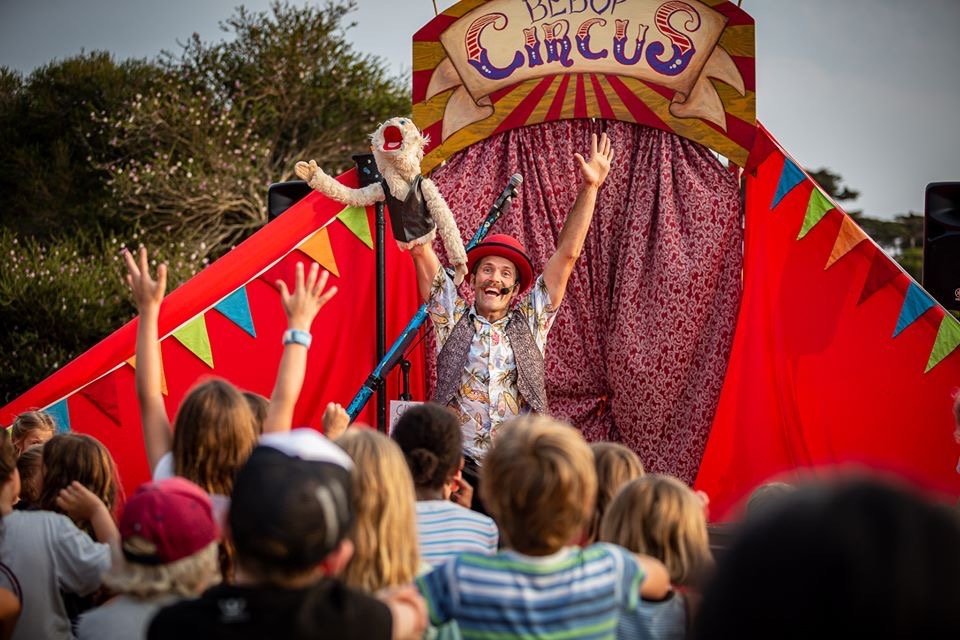 Circus performer in colorful clothing and red hat engaging with children at a circus tent.