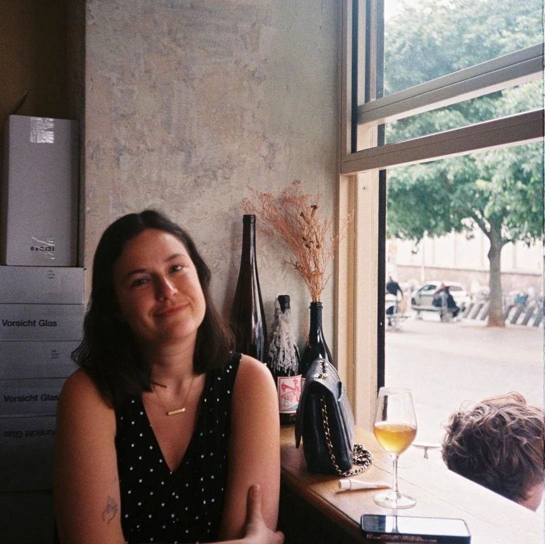 A woman with dark hair sitting at a table in a cafe, smiling slightly, with a glass of white wine and a black purse on the table, next to decorative bottles and dried flowers near a large open window showing an outdoor street scene.