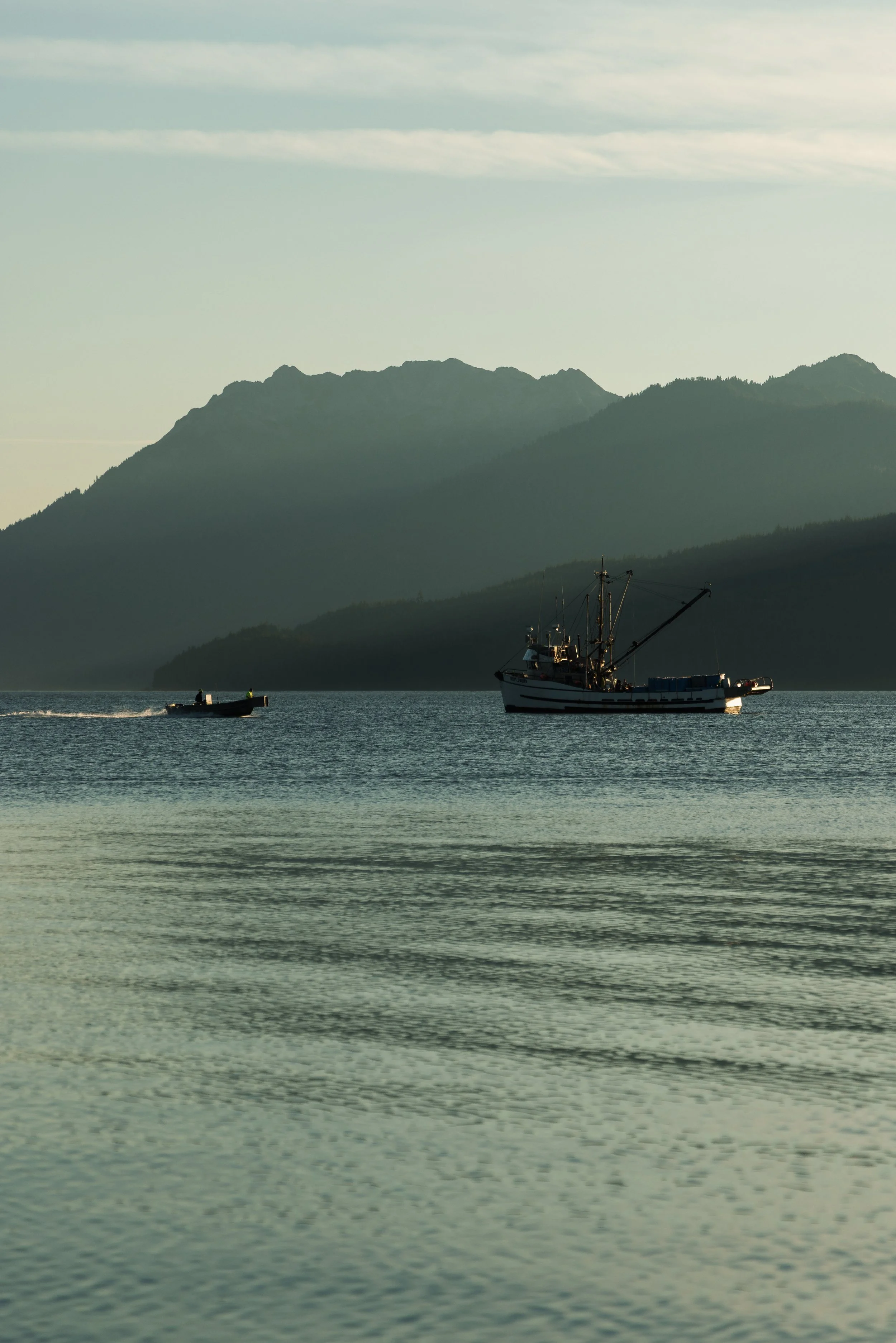 A boat with a crane on it sailing on a body of water with mountains in the background and a partly cloudy sky.