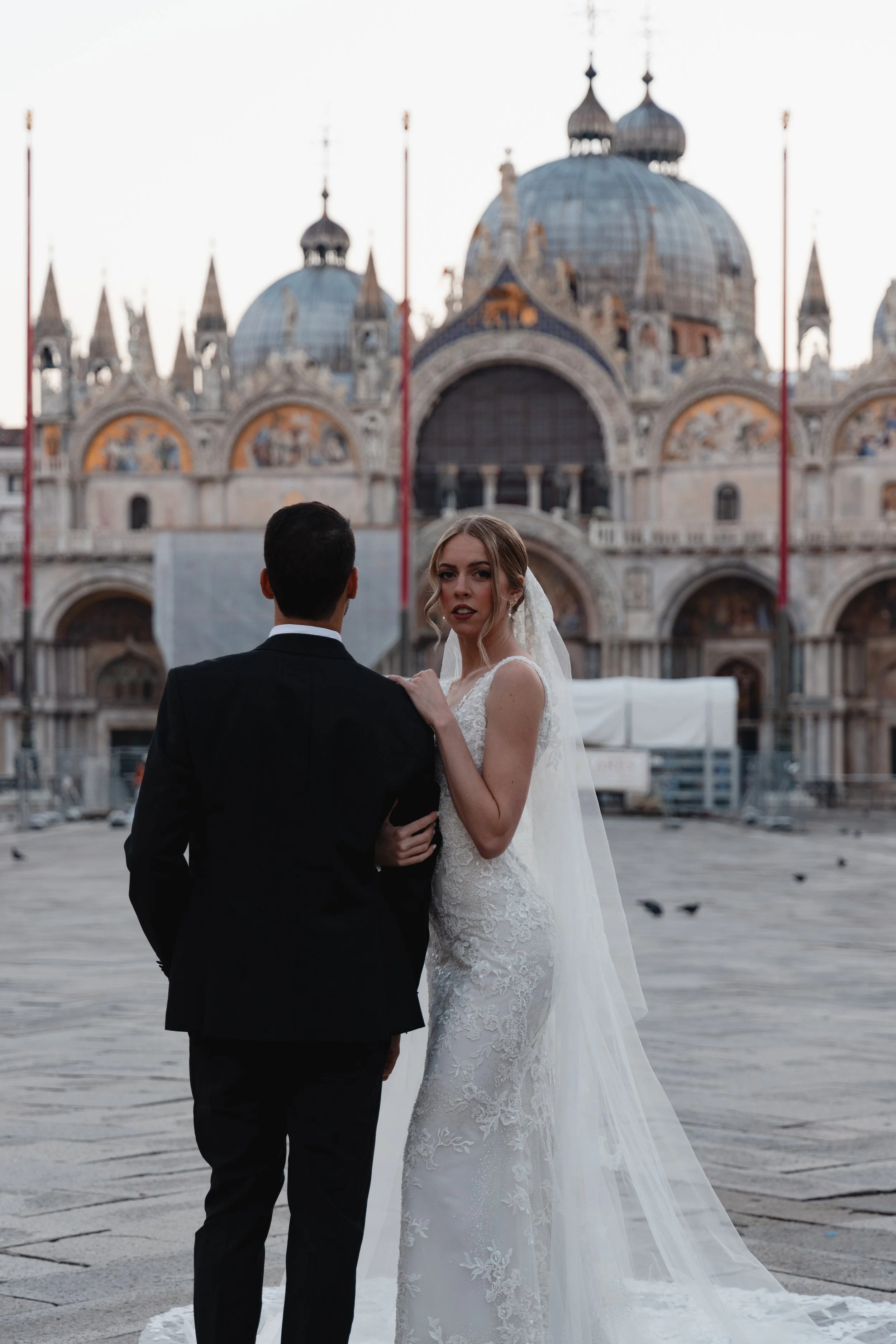 A bride and groom in wedding attire standing in front of St. Mark's Basilica in Venice, Italy, during sunset.