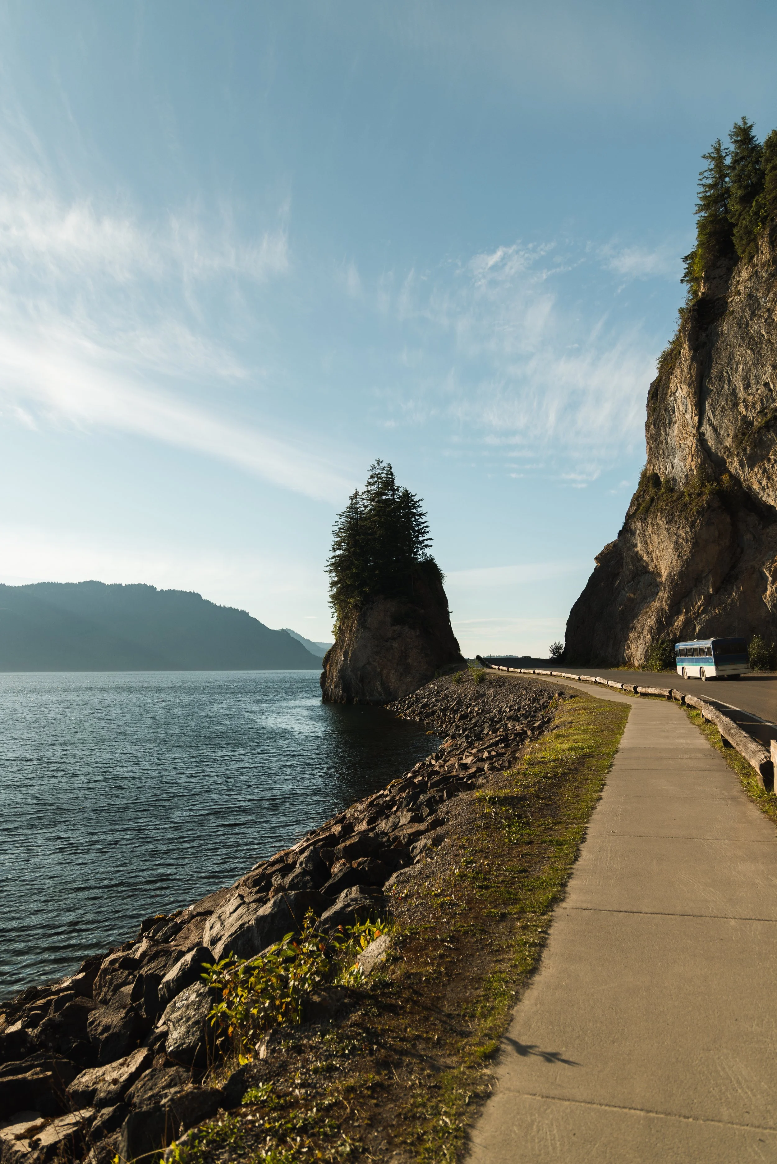 A scenic lakeside view with a paved walkway along the water, rocky shoreline, tall trees on a cliff, and distant mountains under a partly cloudy sky.