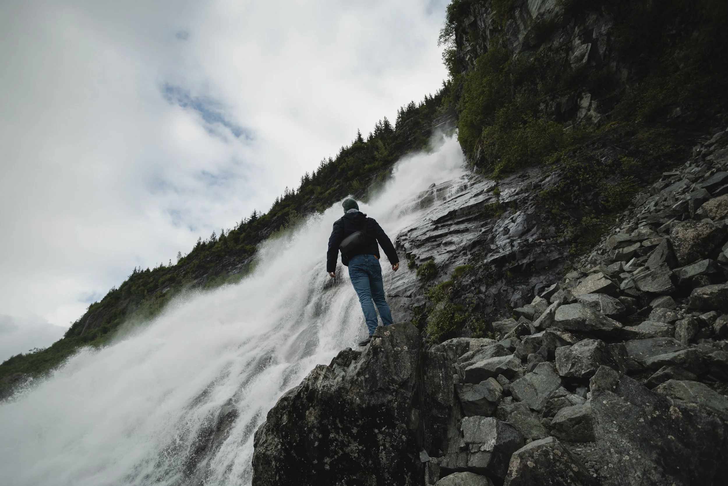 Person hiking on rocky terrain near a cascading waterfall on a mountain slope with trees and cloudy sky in the background.