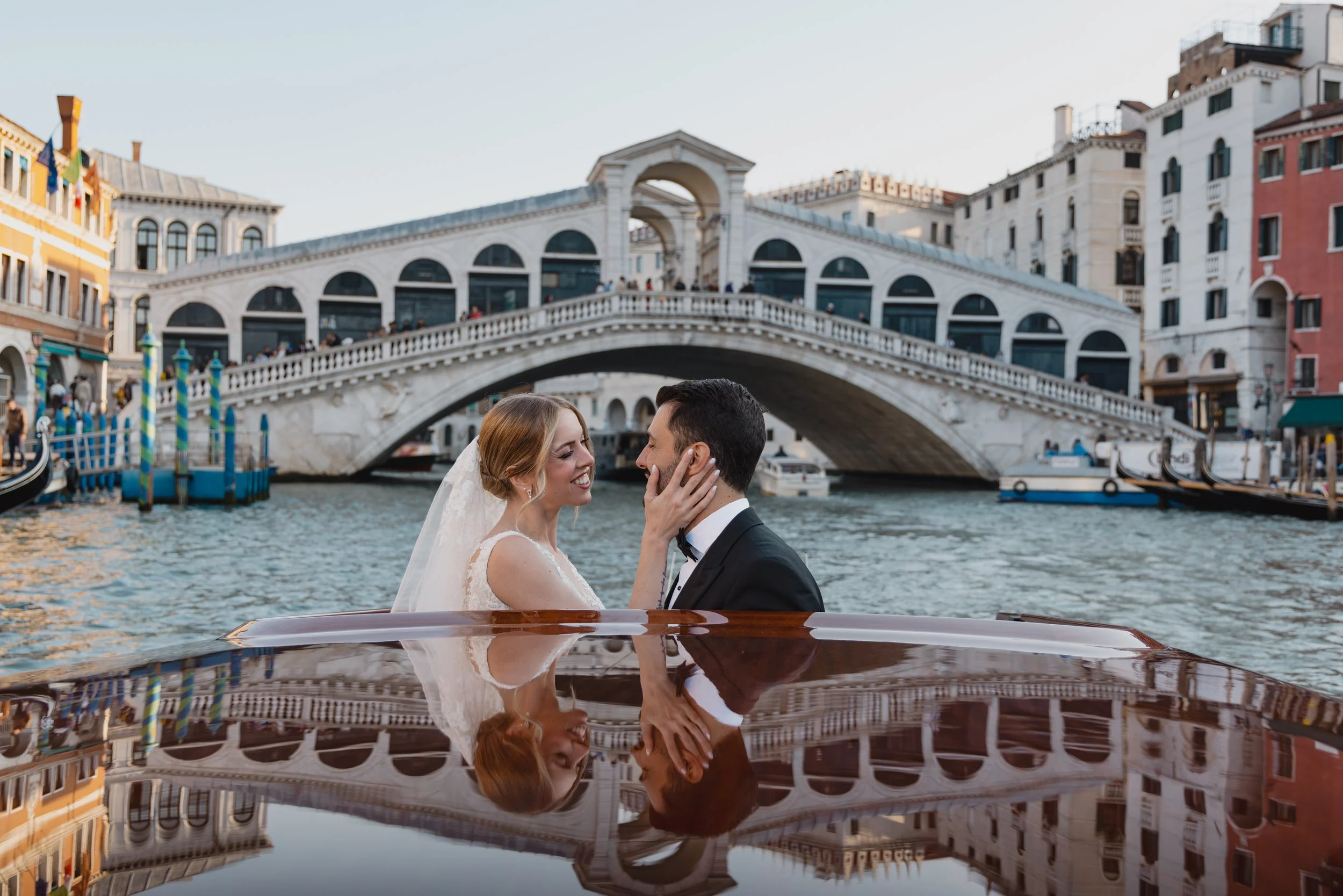 A bride and groom in wedding attire on a boat on a canal in Venice, Italy, with the Rialto Bridge and colorful buildings in the background.