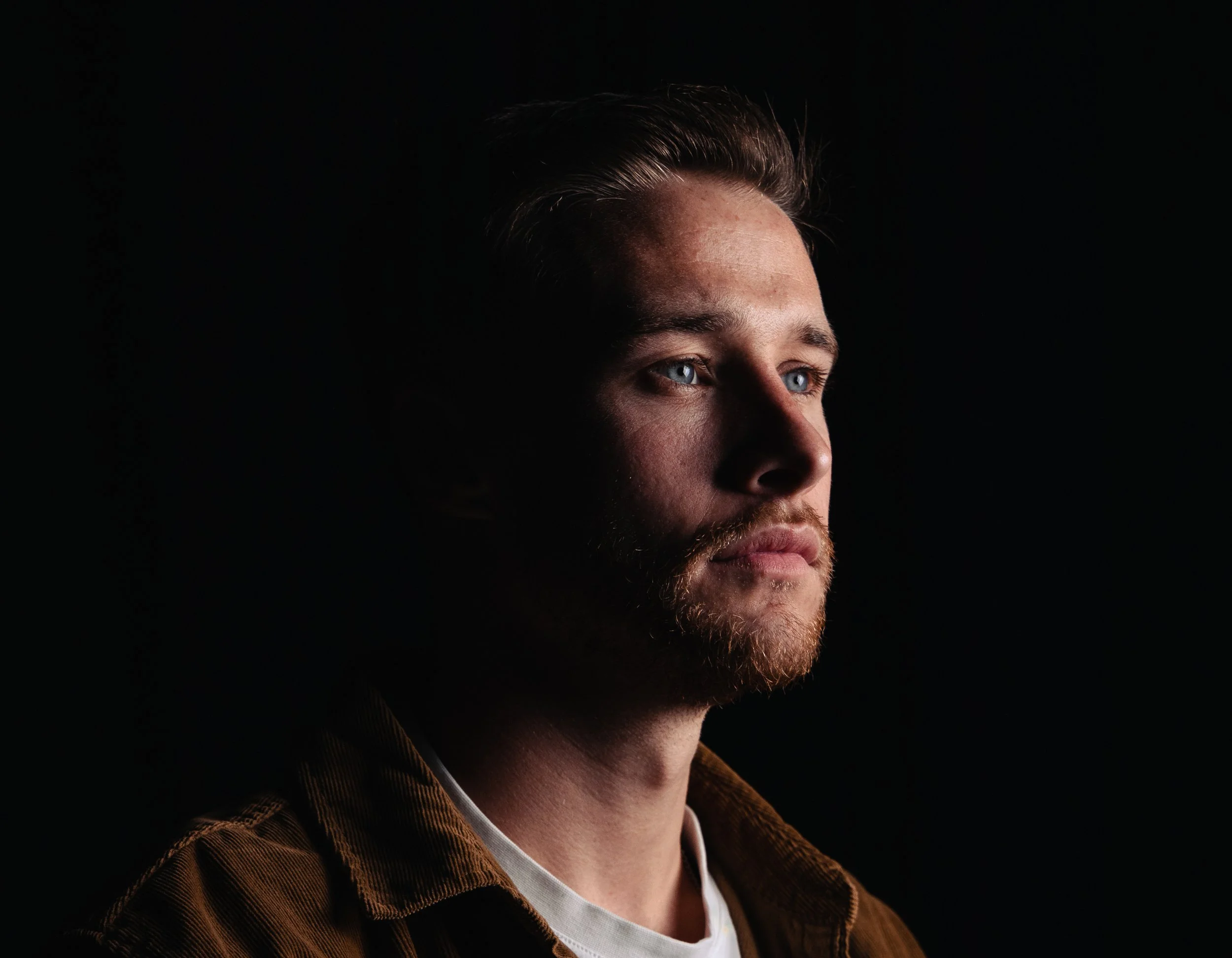 Close-up portrait of a man with blue eyes, brown hair, and a beard, wearing a brown jacket and white shirt, against a dark background.