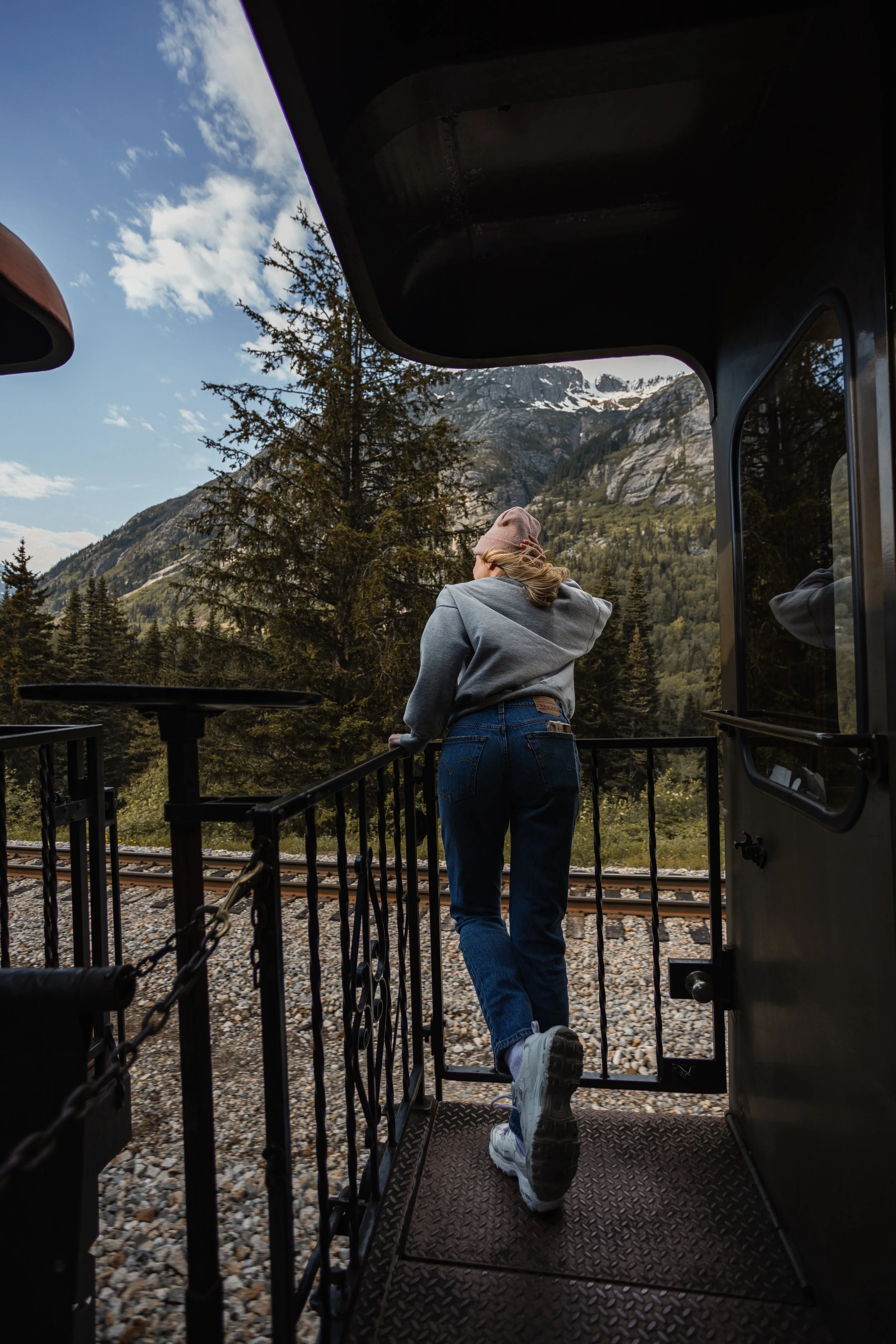 A person wearing a gray hoodie, blue jeans, and white sneakers walks out of a train onto a scenic mountain landscape with trees and snow-capped peaks in the background.