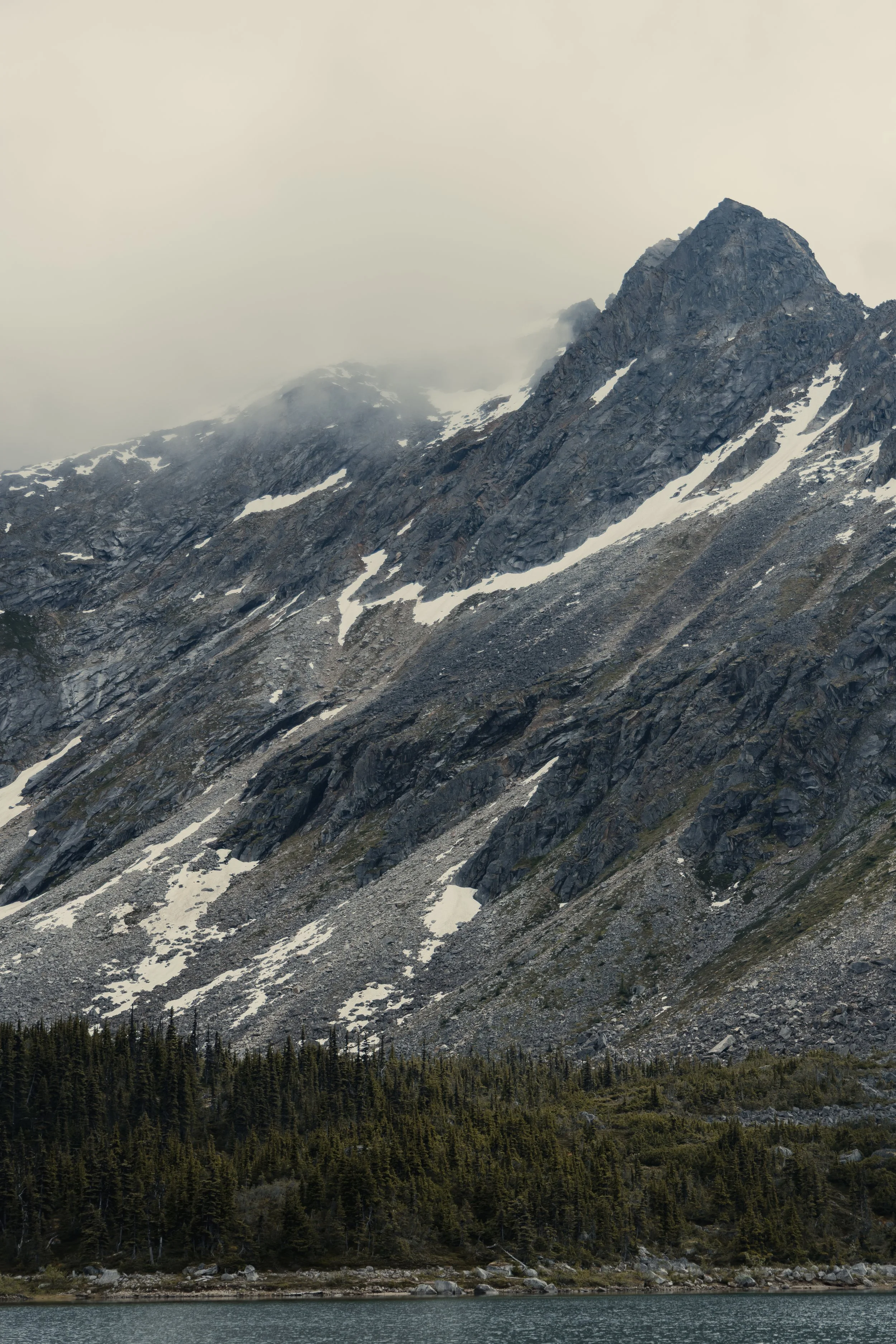 Mountain landscape with rocky peaks, patches of snow, a forest at the base, and a lake in the foreground under a cloudy sky.