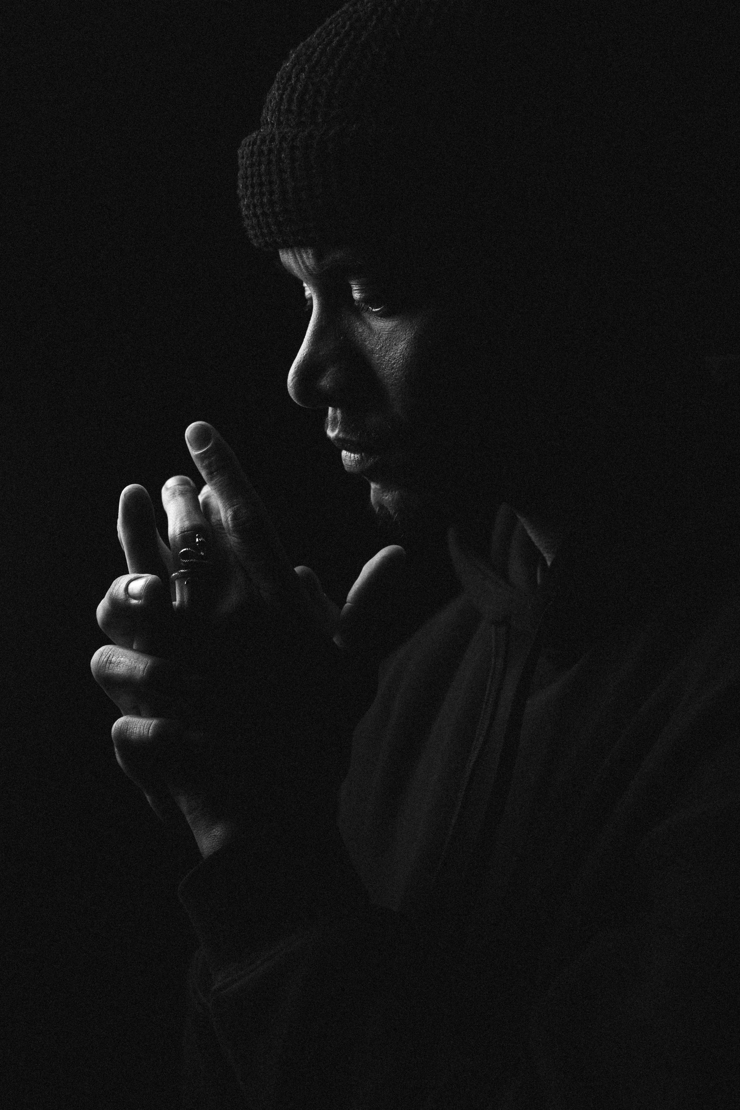 Black and white portrait of a person wearing a knit beanie, deeply in thought, with their hands clasped together near their face.